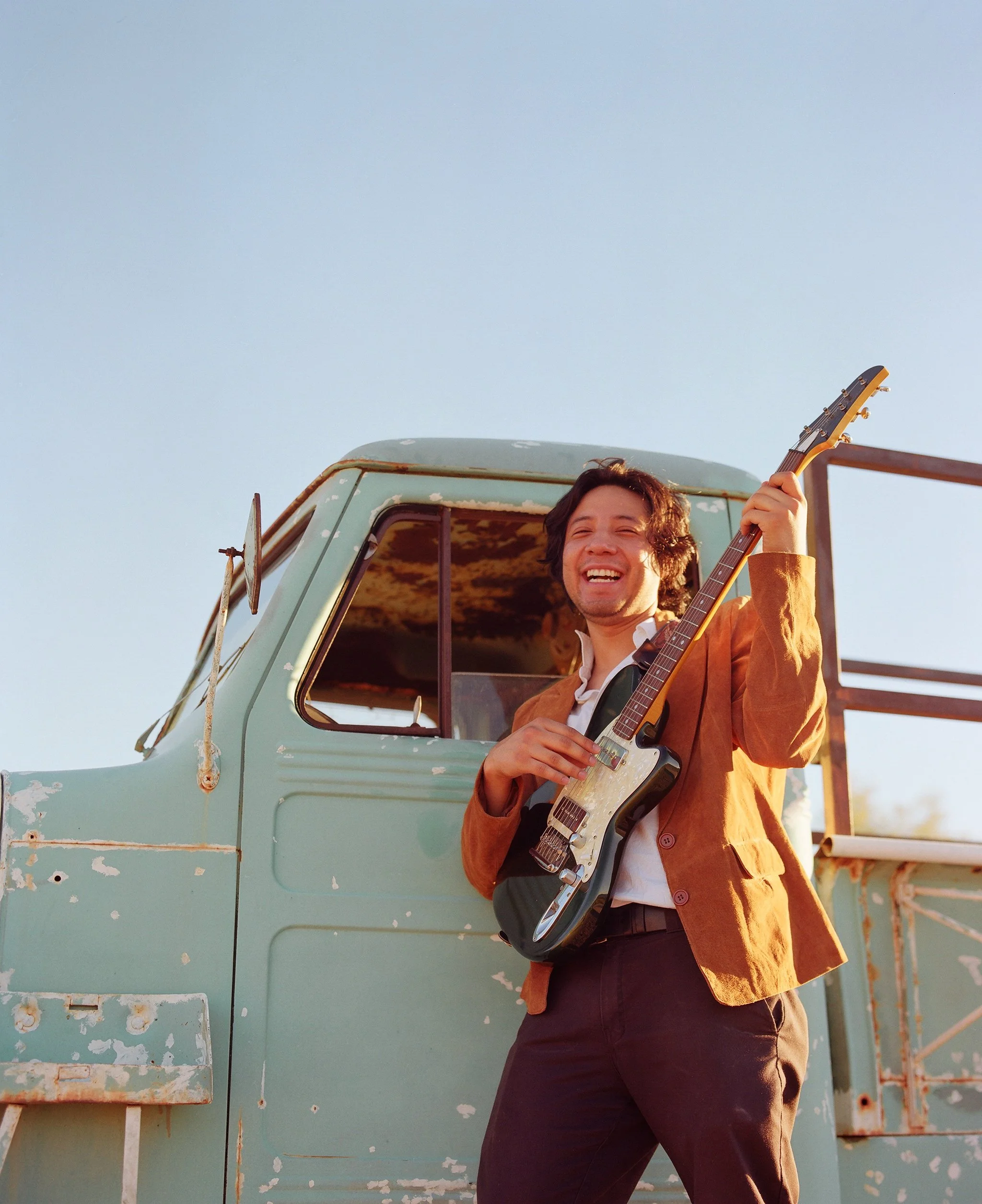 A person smiling while holding an electric guitar, standing next to an old, weathered green truck with peeling paint and rust, outdoor setting with clear blue sky.