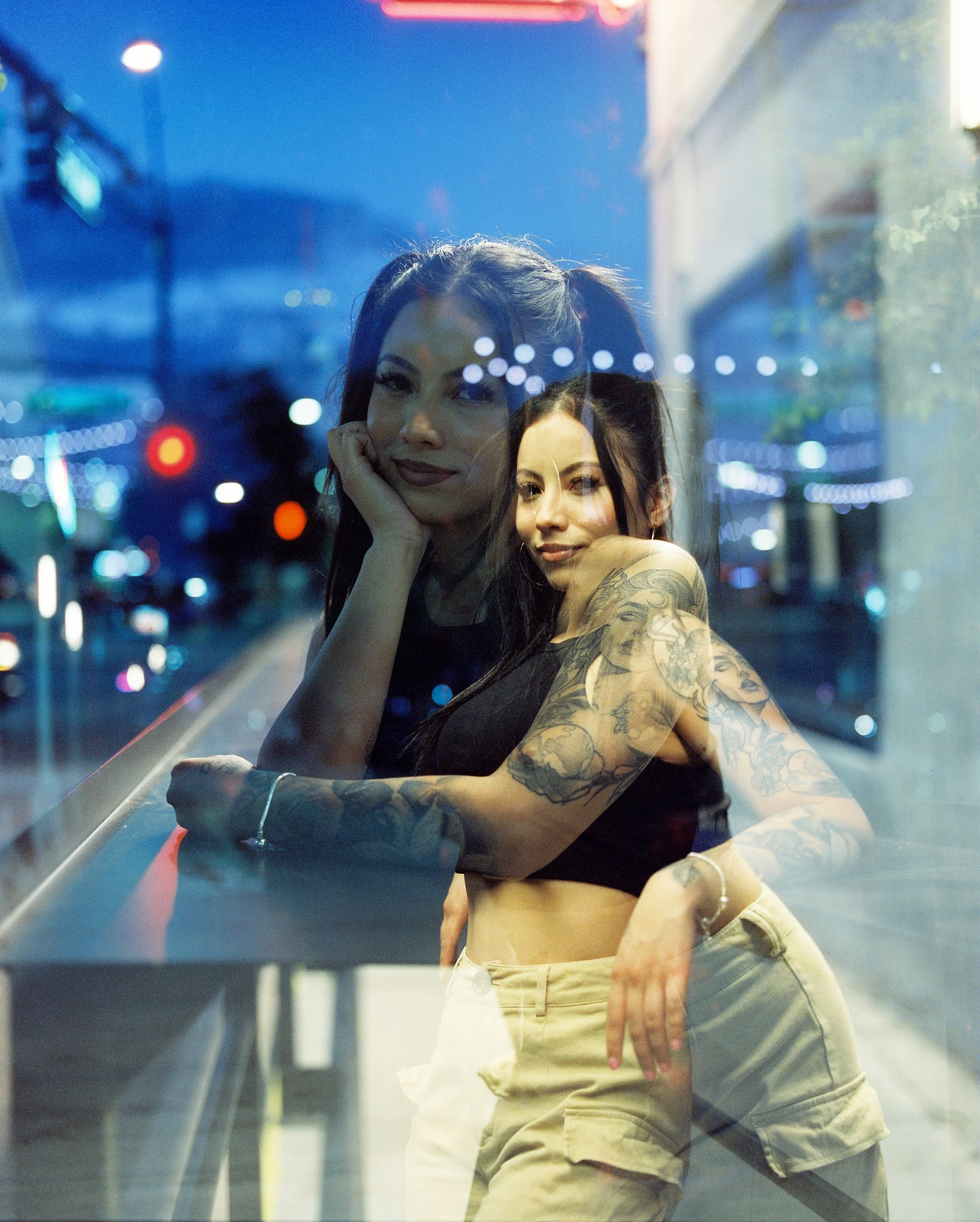 A young woman with tattoos on her arms and face, wearing a black top and beige shorts, standing behind a reflective window at night with blurred city lights and street signs in the background.