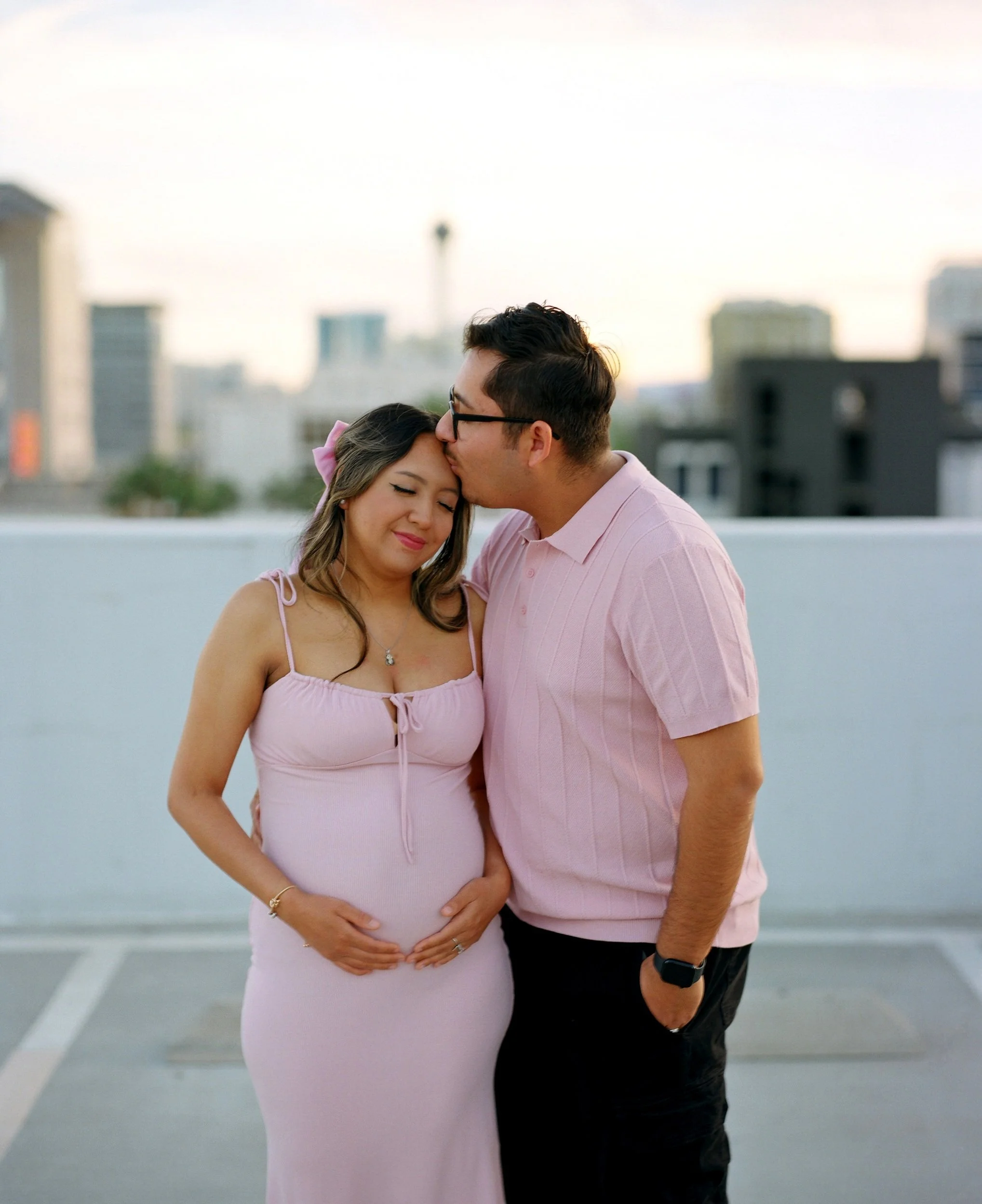 A pregnant woman in a pink dress with a pink bow in her hair, and a man in a pink shirt sharing a moment with her, on a rooftop with city buildings in the background.