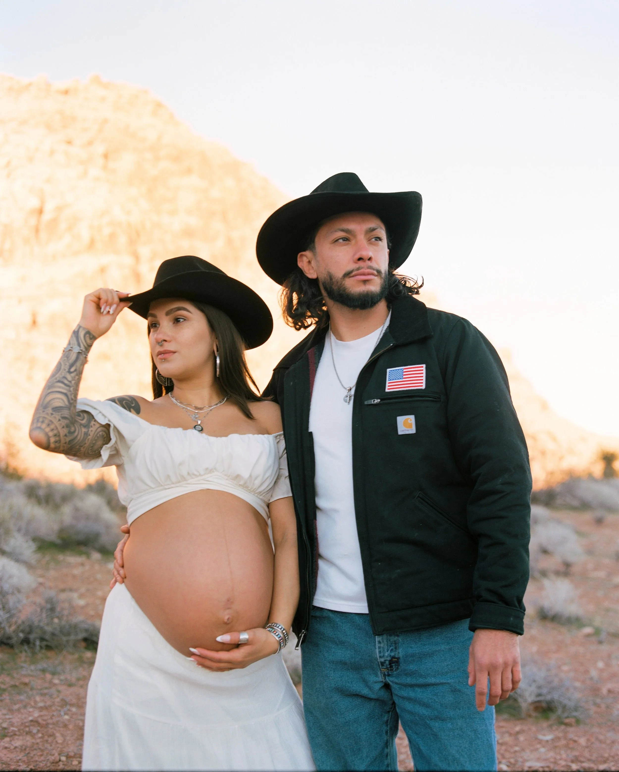 A pregnant woman with tattoos on her arm, wearing a white dress and a black hat, standing outdoors with a man in a black jacket and a black hat. The woman is touching her hat with one hand and her belly with the other, while the man gazes into the di