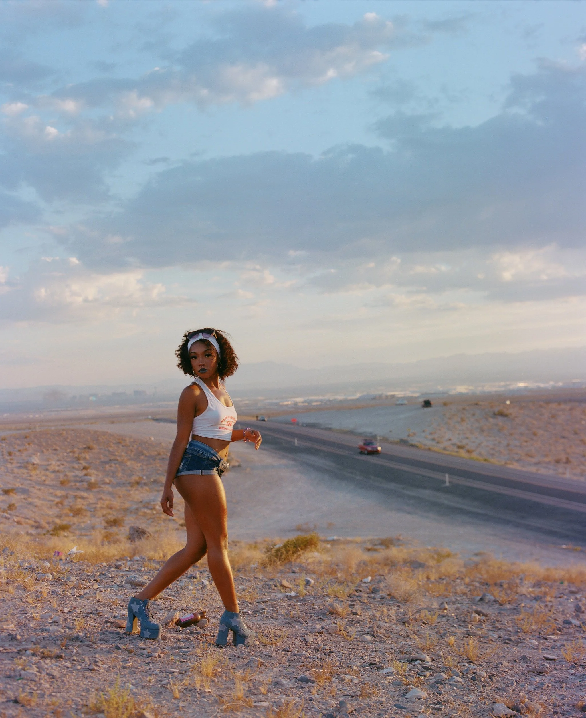 A woman with curly hair wearing star-patterned high-heeled boots, a white tank top, and denim shorts walking on a gravel path in a desert-like area with a road and cars in the background under a cloudy sky.