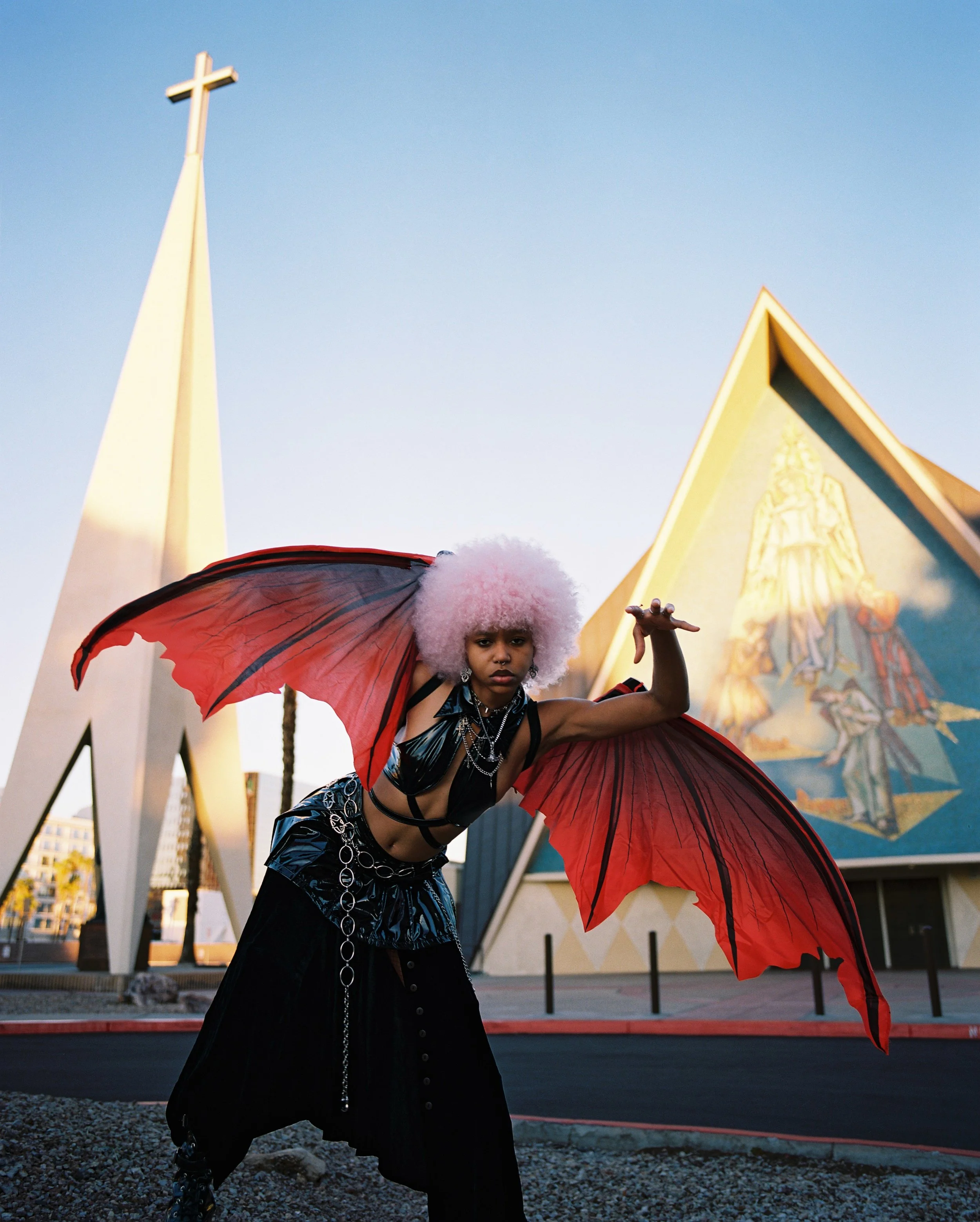 A person with pink curly hair, wearing dark clothing with chains, holding a large red umbrella with black lining, standing in front of a modern building with a mural and a church steeple with a cross, in an urban setting during sunset.