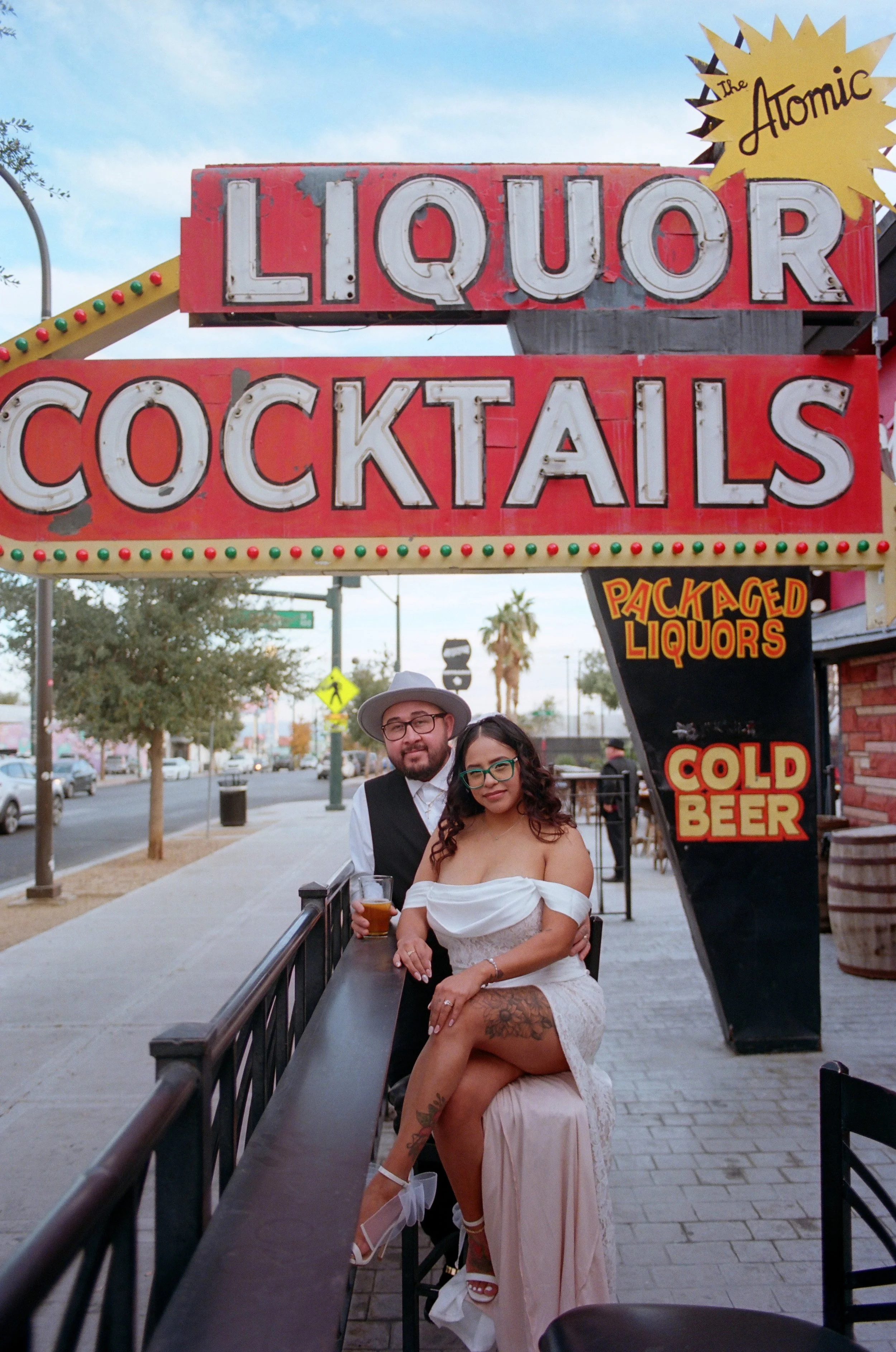 A couple standing at an outdoor bar, with the woman sitting on a man's lap, holding a drink in her hand, in front of a neon sign that says 'Liquor Cocktails' and other signs for packaged liquors, cold beer, and the 'Atomic' sign, with street and trees in the background.