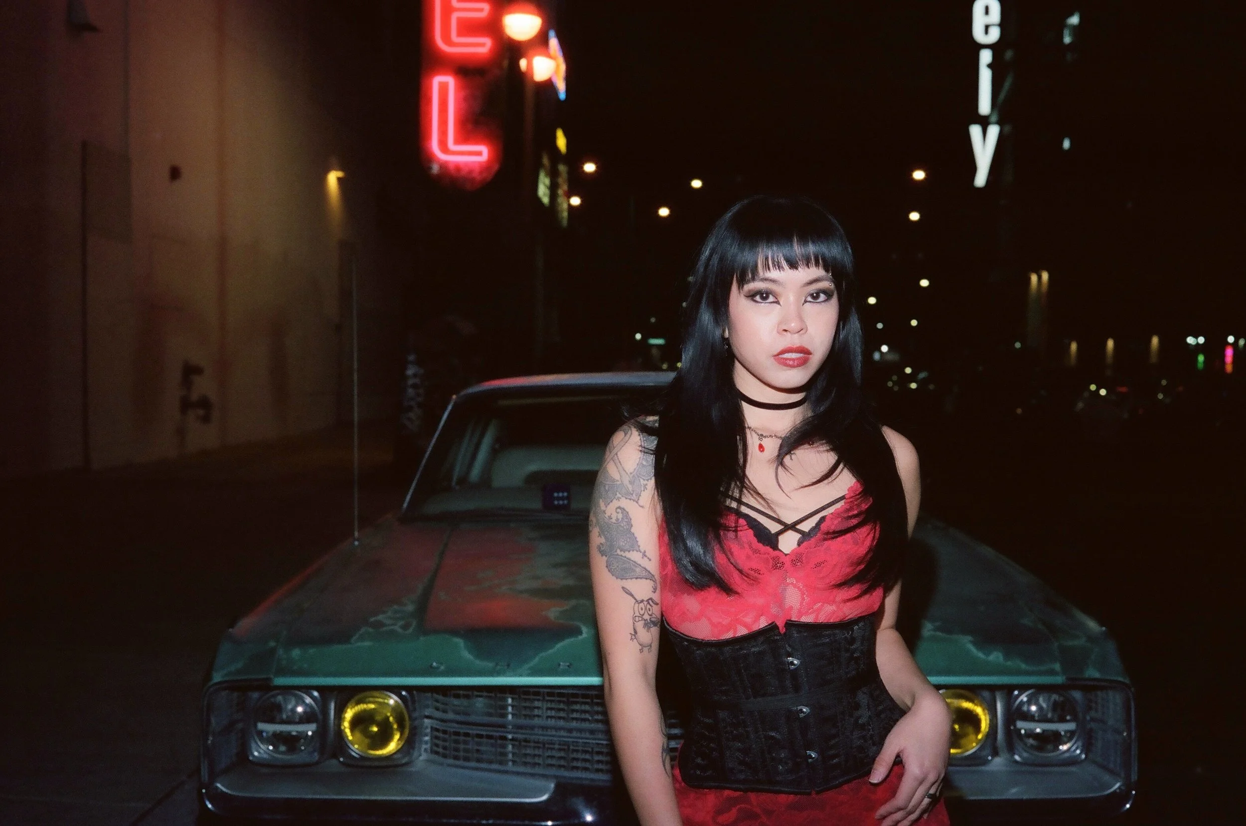 A woman with black hair, tattoos, and red lipstick standing in front of a vintage car at night, with neon and city lights in the background.