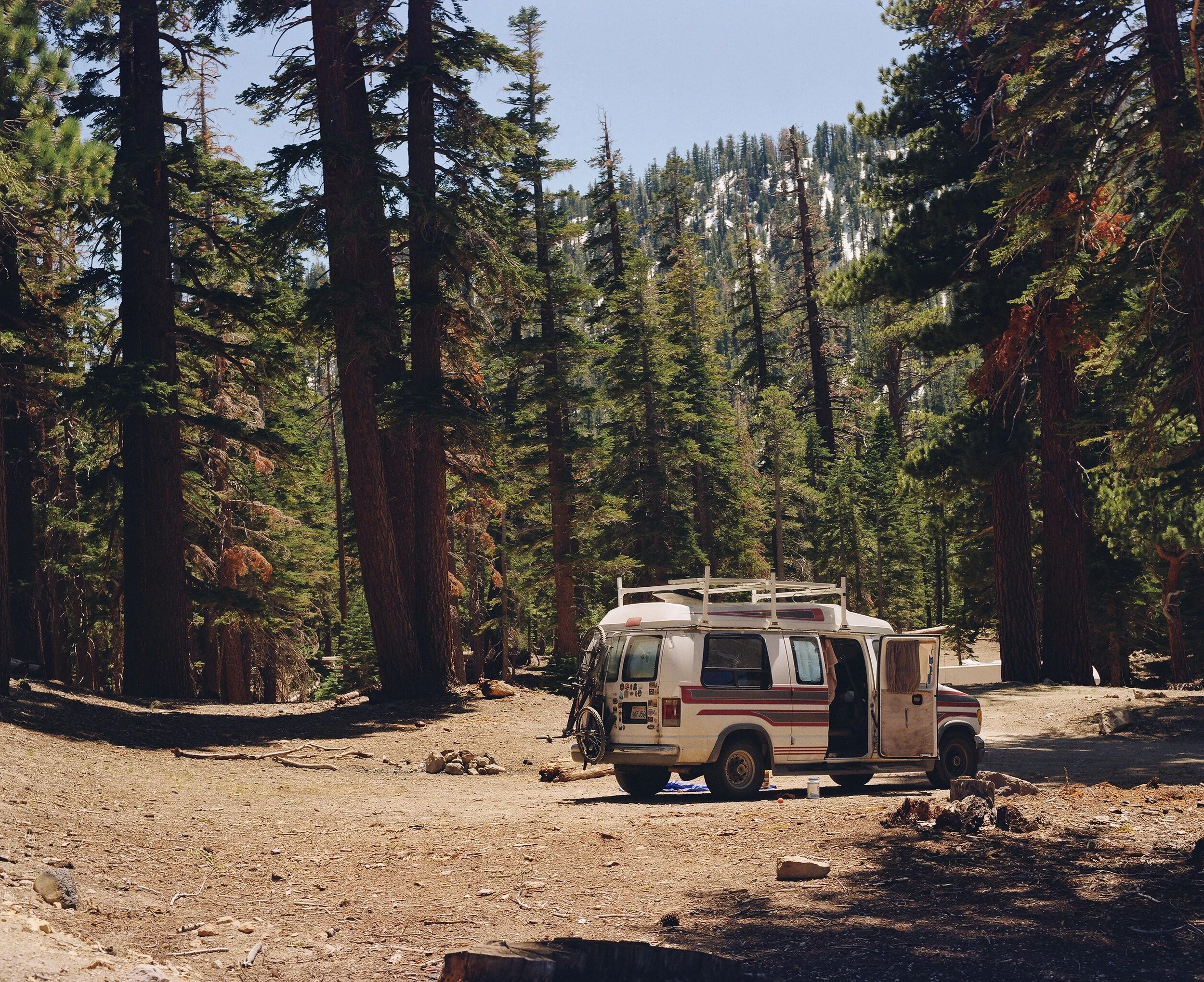 A camping site in a forest with tall pine trees and a snowy mountain in the background. There is an RV and a bicycle parked near the trees, and the ground is dirt with some rocks and a tree stump in the foreground.