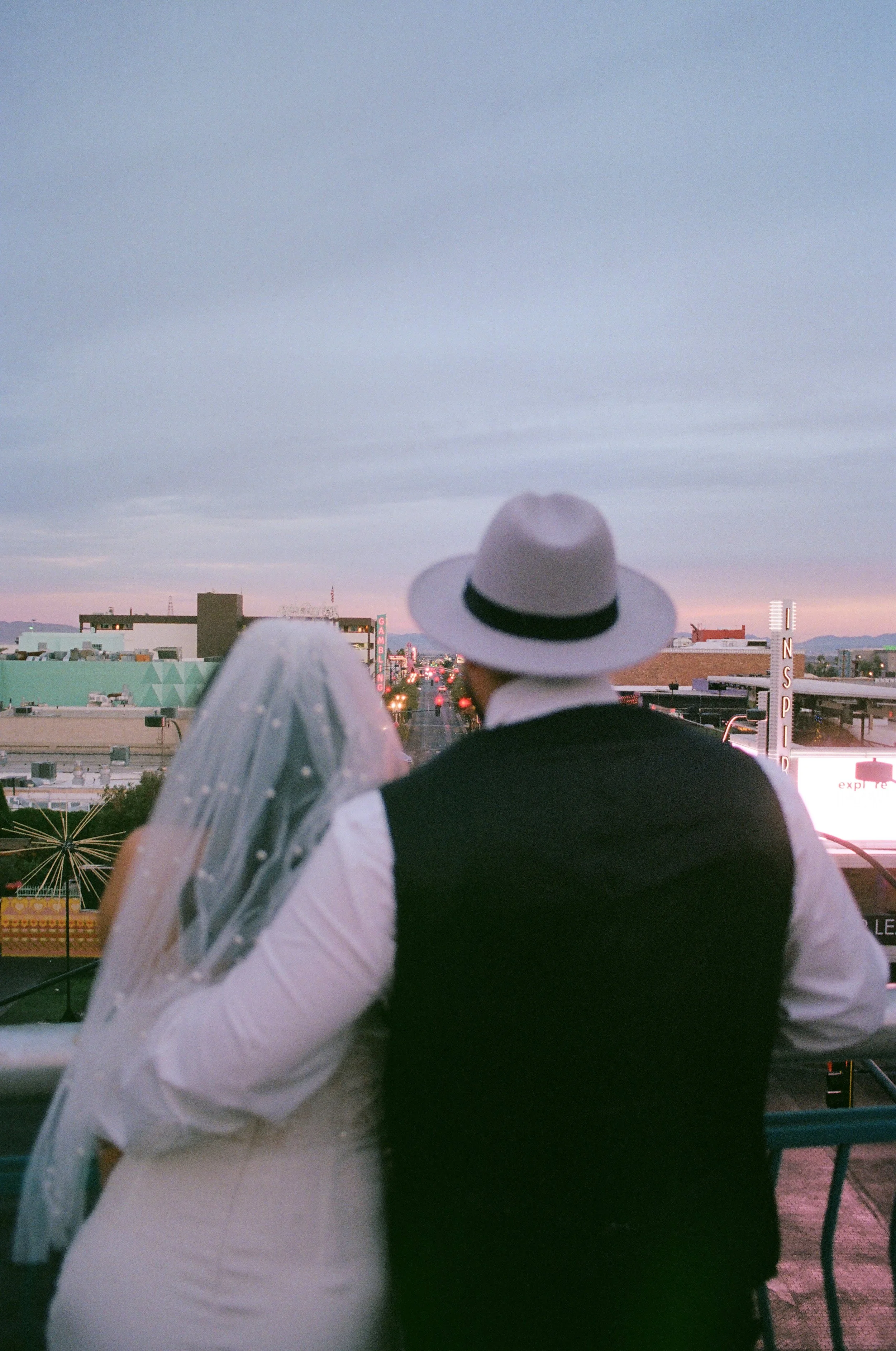 A man dressed in black and white, wearing a white wide-brimmed hat, is looking over a cityscape during dusk, with cloudy skies and neon signs in the background.