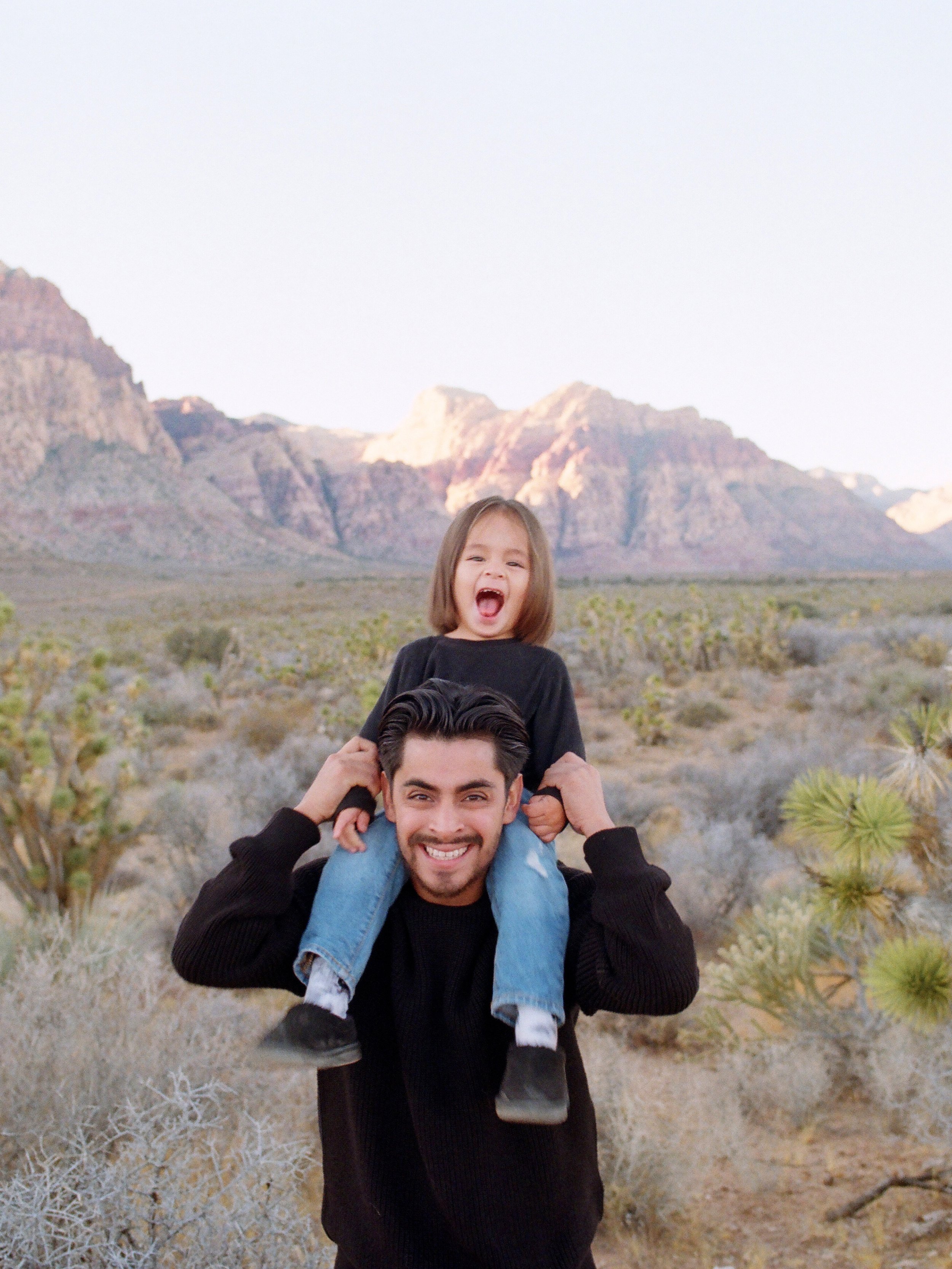 A man playing with a young boy on his shoulders in a desert landscape with mountains in the background.