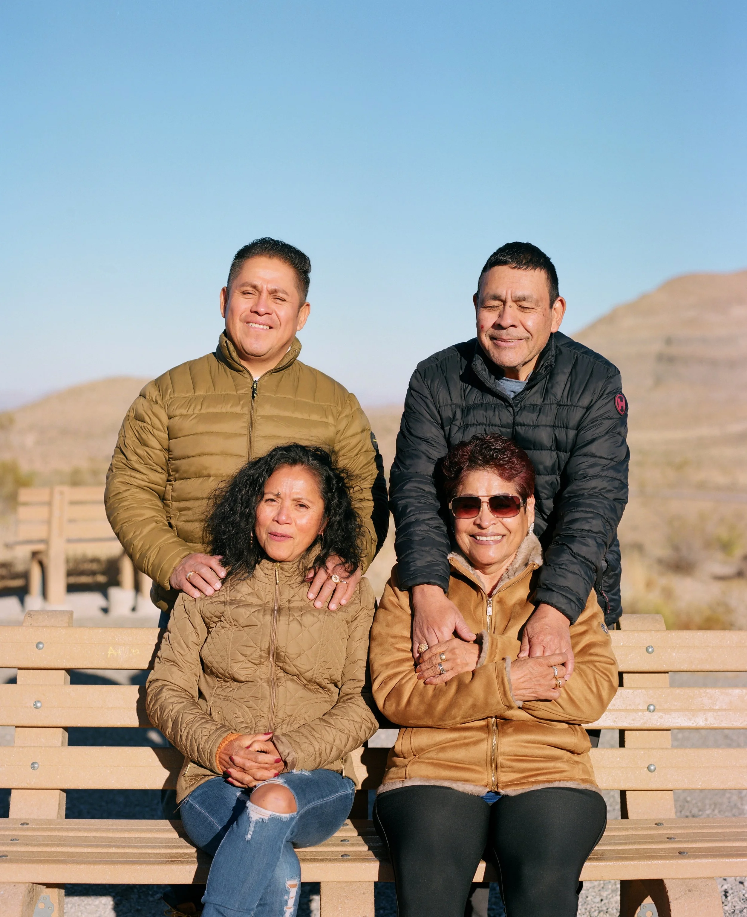 A group of five diverse people sitting on a wooden bench outdoors in a sunny, mountainous area. Two women are seated front, with two men standing behind them and another person’s head partially visible at the bottom. They are smiling, with some weari