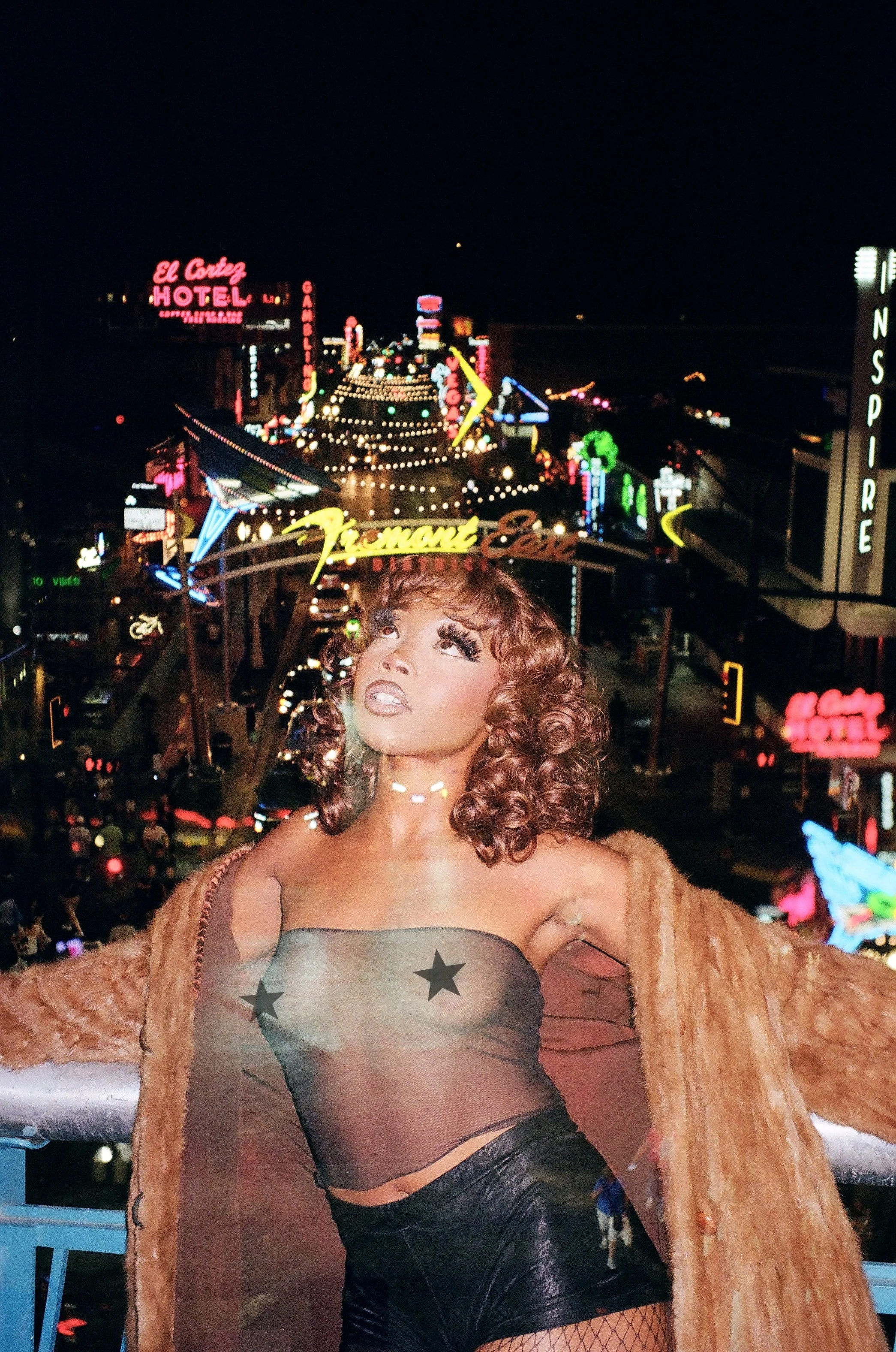 Nighttime scene of a neon-lit street with signs for hotels, theaters, and restaurants, with a woman in the foreground wearing a furry coat, sheer top with stars, and fishnet stockings.