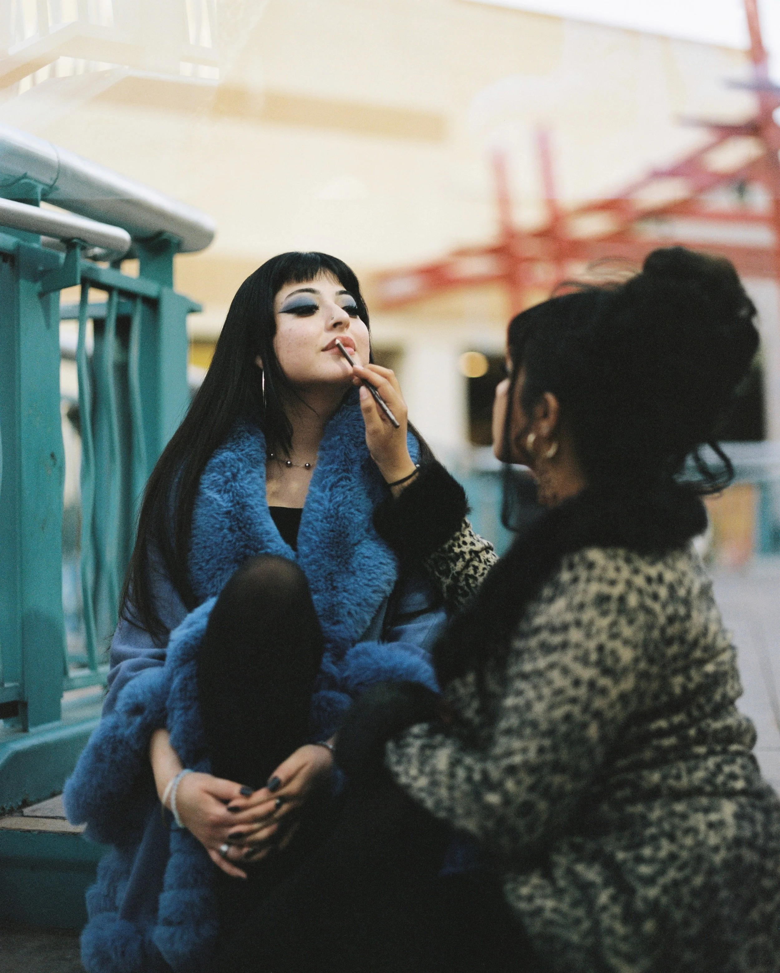 Two women are talking near a teal railing in an indoor setting. The woman with long dark hair is applying makeup to the other woman, who has short dark hair and is wearing a leopard-patterned coat.