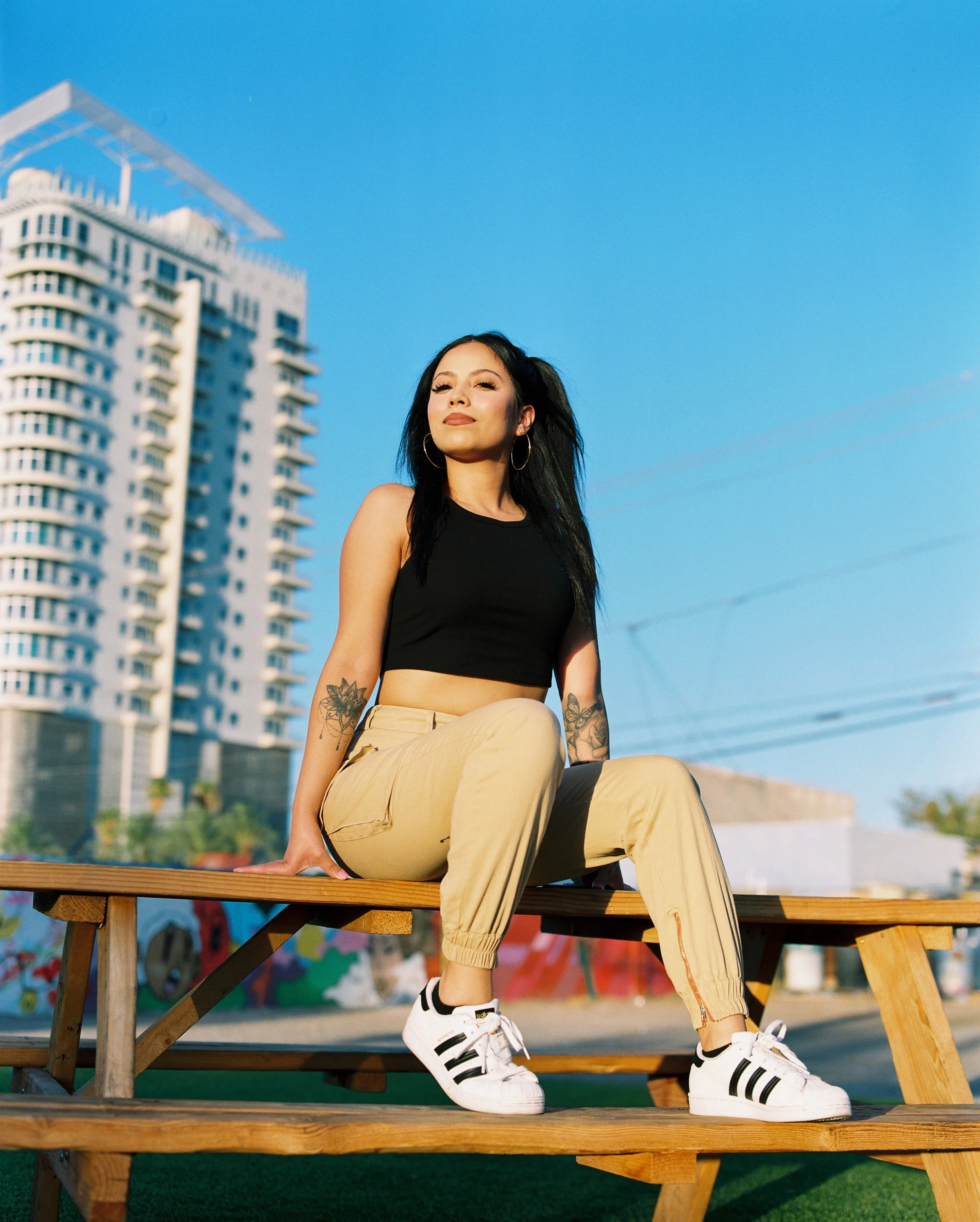 A young woman with black hair, hoop earrings, and tattoos on her arms, sitting on a wooden table outdoors against a cityscape background with tall buildings and a clear blue sky. She is dressed in a black crop top, beige joggers, and white sneakers with black stripes, looking confidently at the camera.