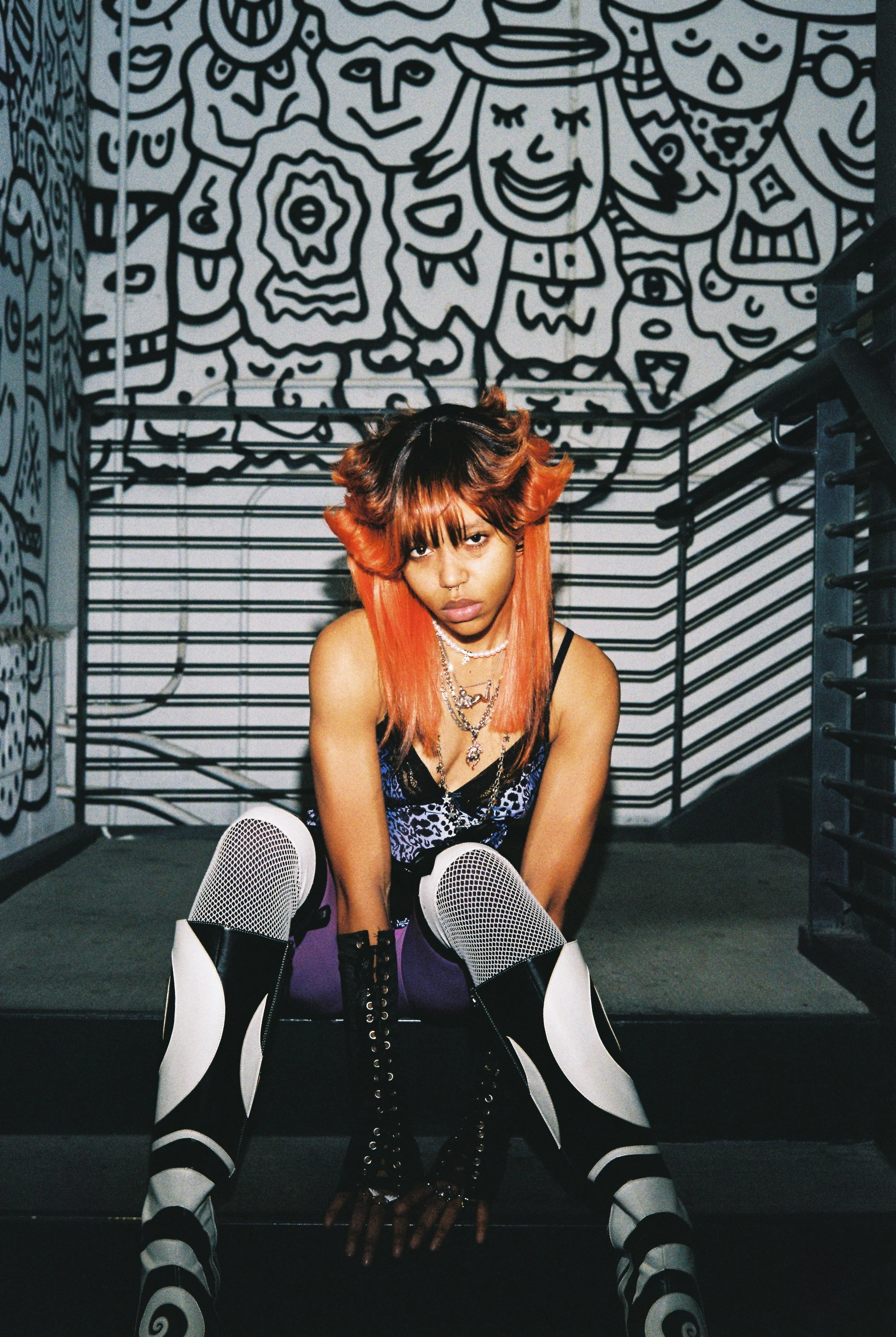 A young woman with orange and black hair sitting on the floor in front of a black-and-white wall with cartoon drawings. She is wearing fishnet stockings, black platform shoes, and layered necklaces, and has a serious expression.