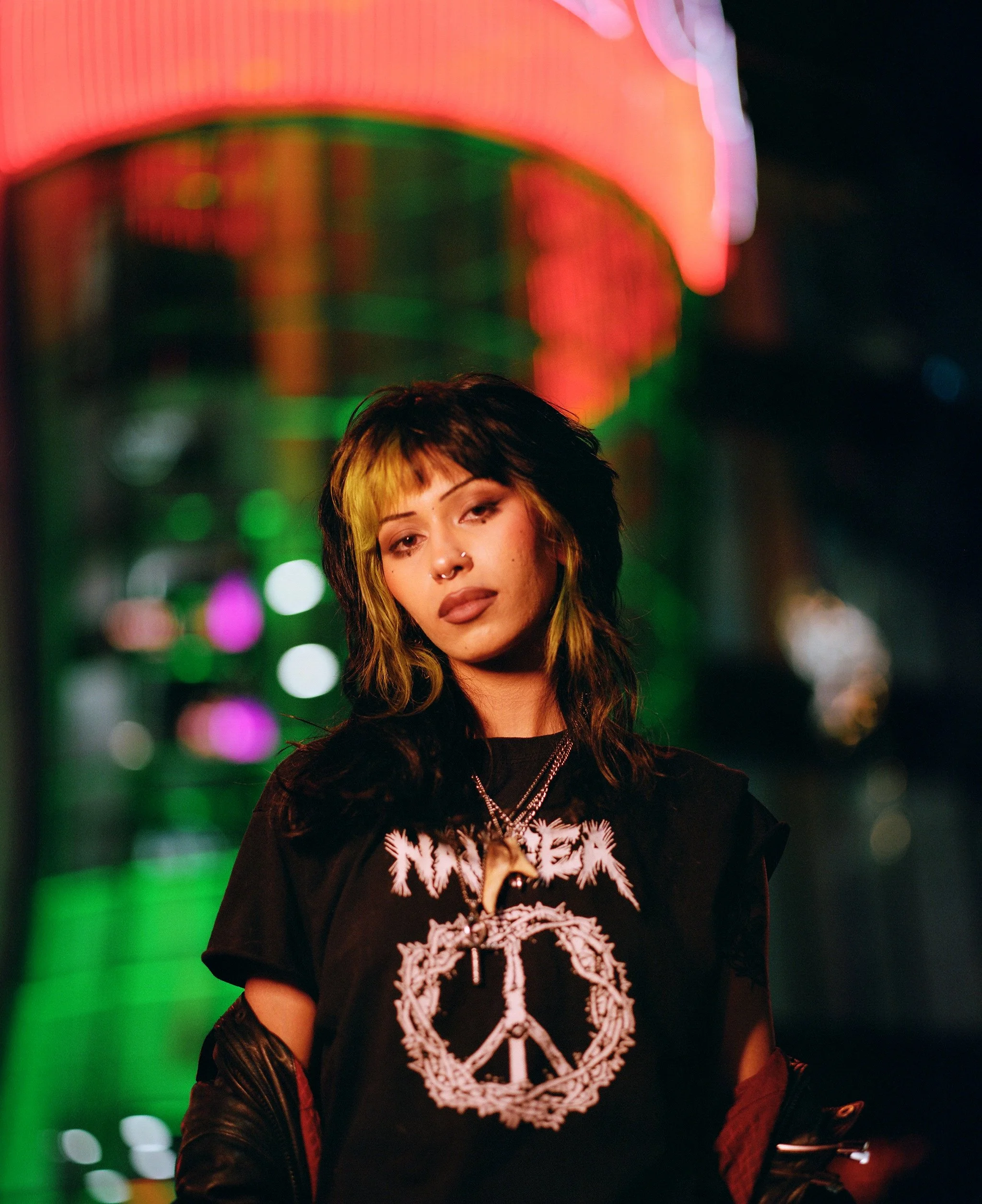 A young woman with black hair and yellow highlights, wearing a black graphic T-shirt with a white dragon design, a black leather jacket, and posing with neon-colored lights and bokeh in the background.