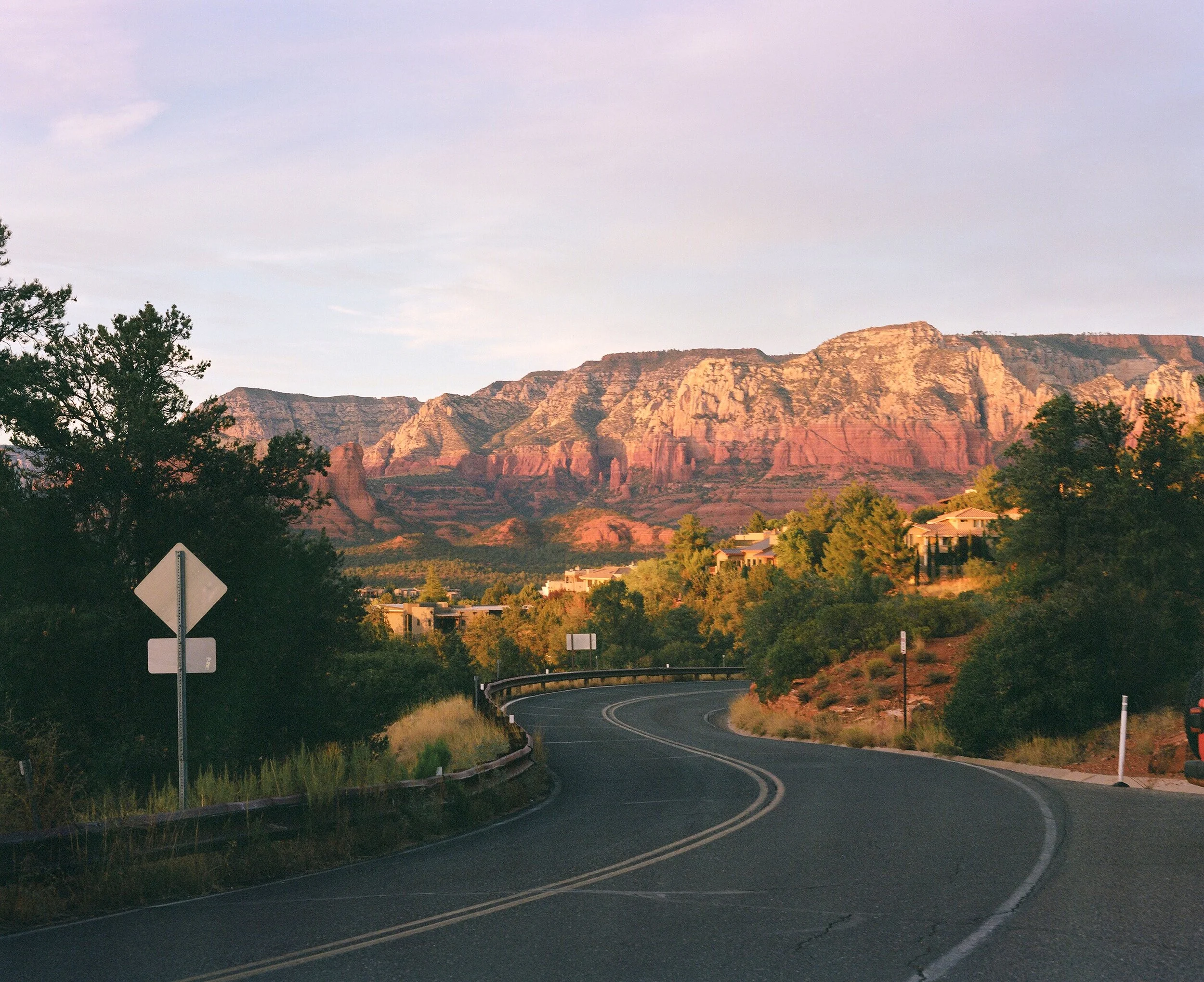 A winding road with double yellow lines in a hilly, desert landscape at sunset. There are some trees, houses, and a sign in the foreground, and red rock formations and mountains in the background.