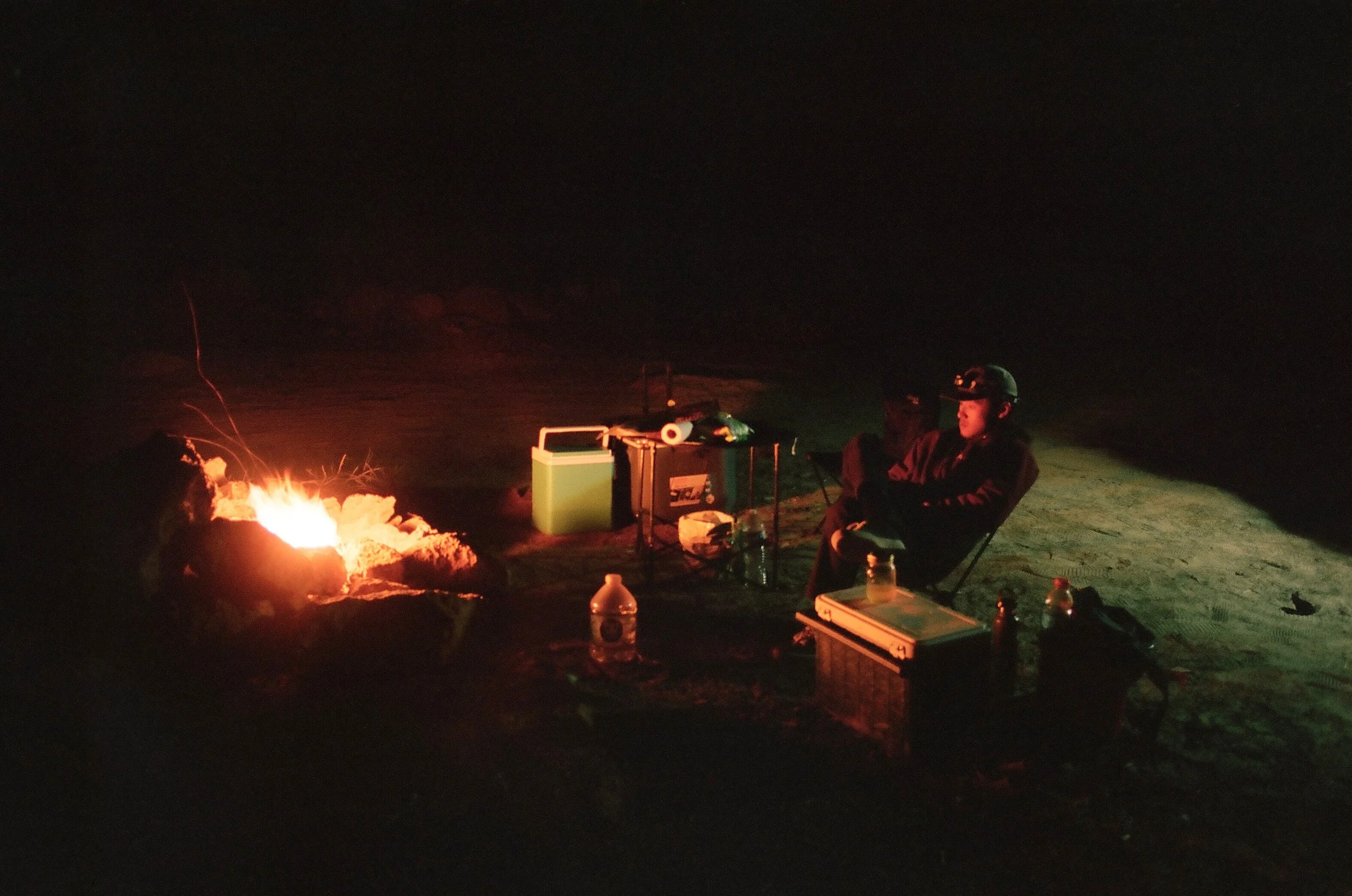 Person sitting by a campfire at night, with camping supplies nearby.