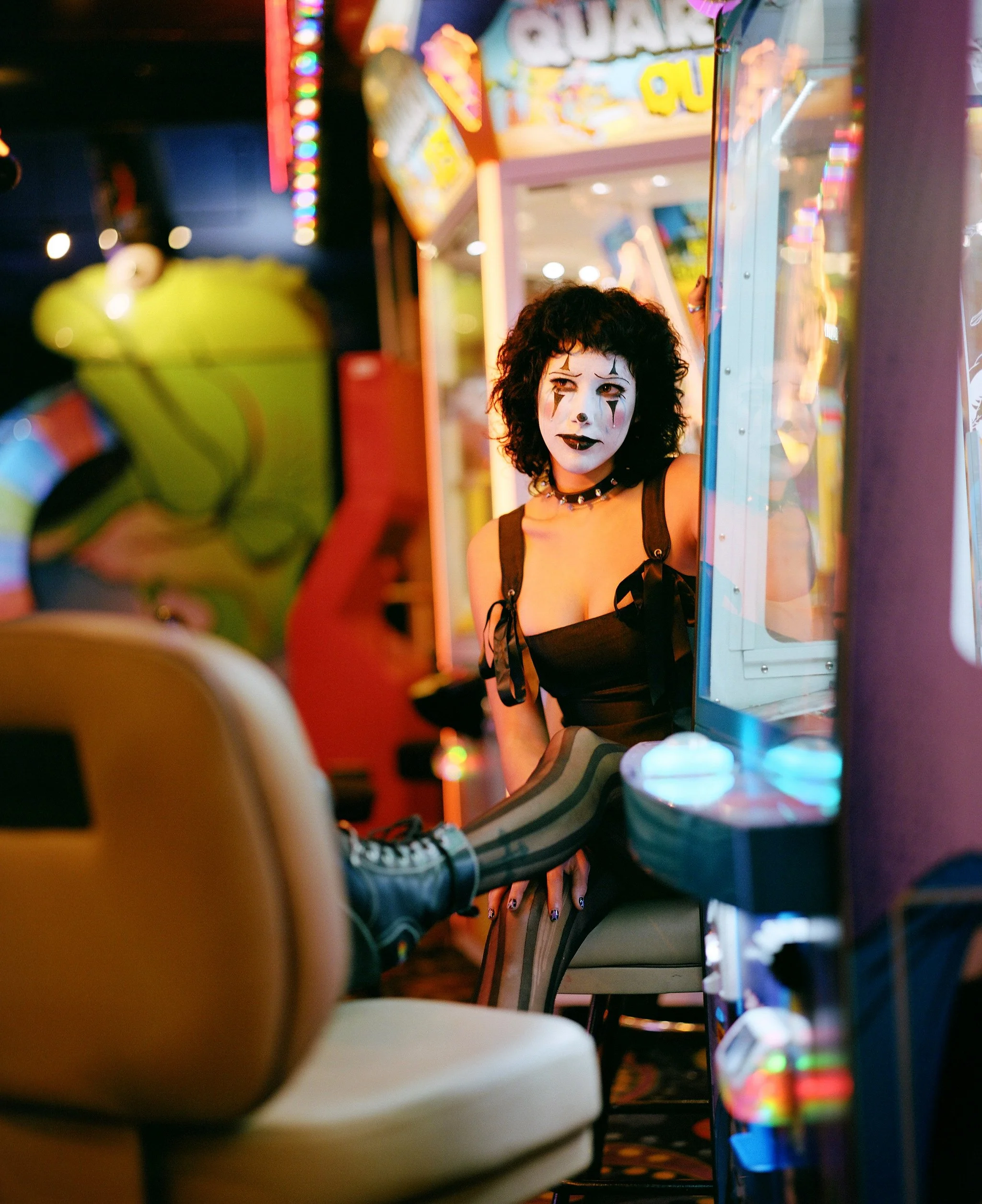 A woman with black curly hair and clown makeup, wearing a black sleeveless top, sitting at a slot machine in an arcade or casino. The background has colorful lights and signs.