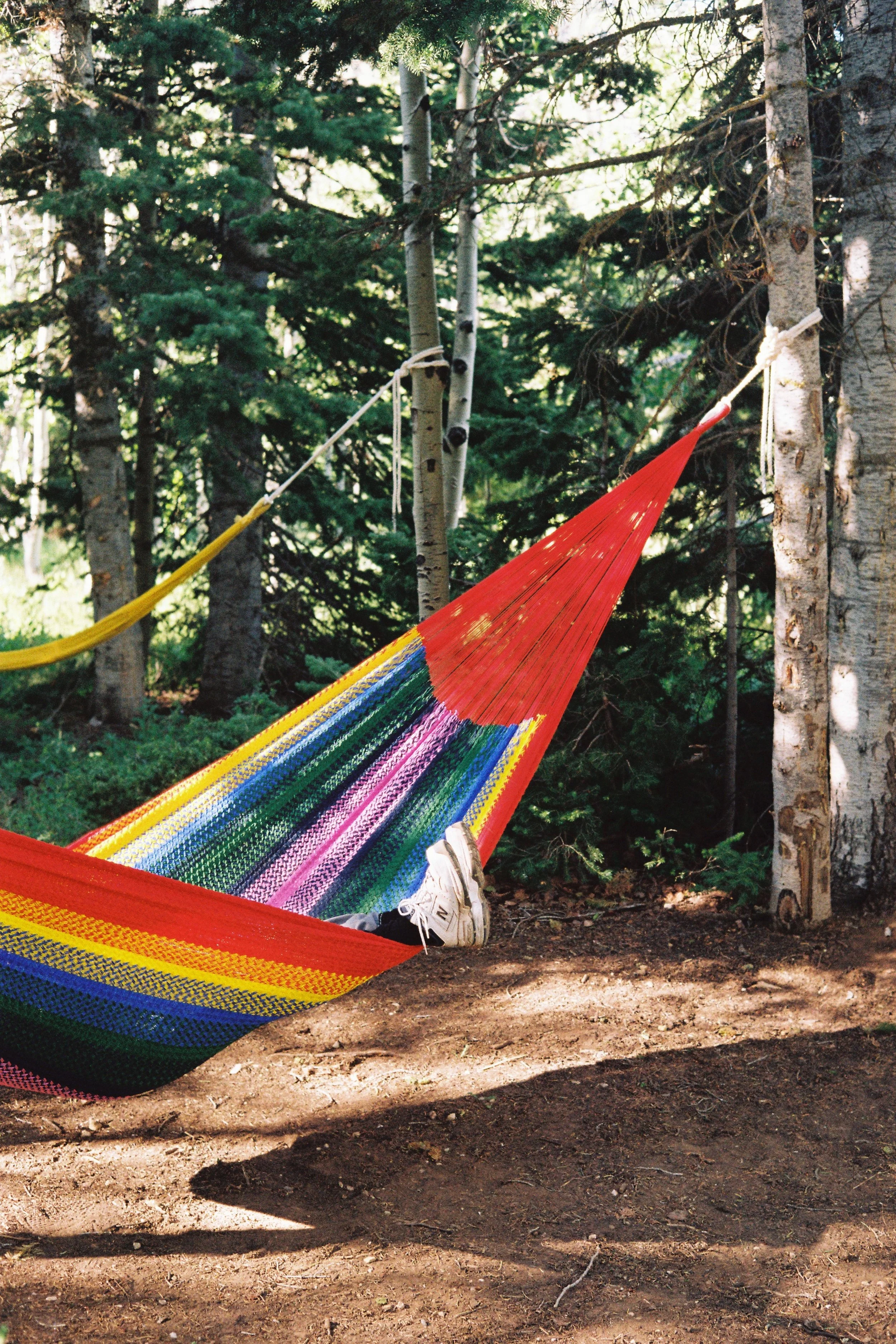 A colorful striped hammock tied between trees in a forest, with a person wearing white sneakers lying inside, with their legs hanging outside the hammock.