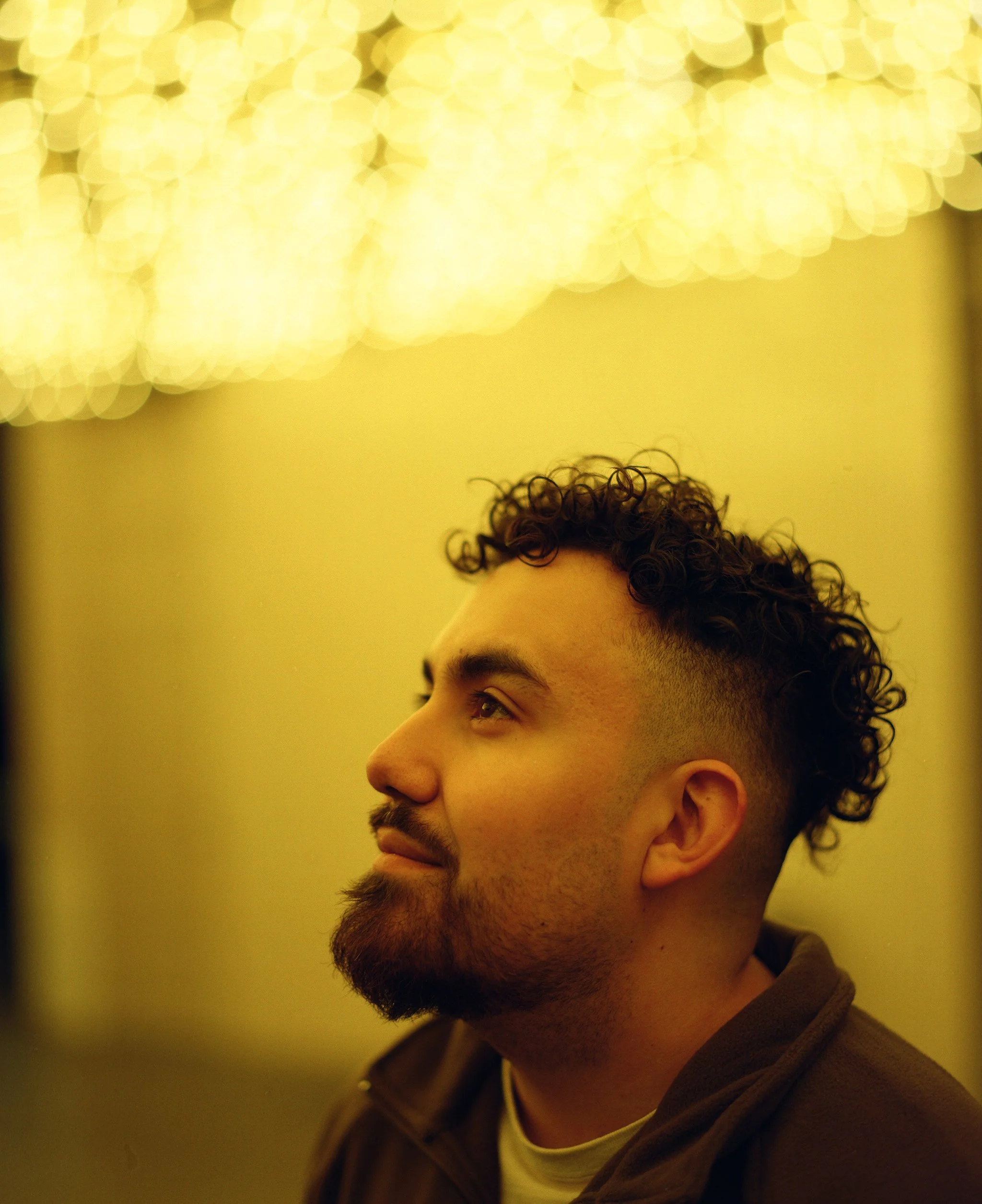 Profile side view of a young man with curly dark hair and a beard, wearing a brown hoodie and a light-colored shirt, against a yellow background with blurred lights.