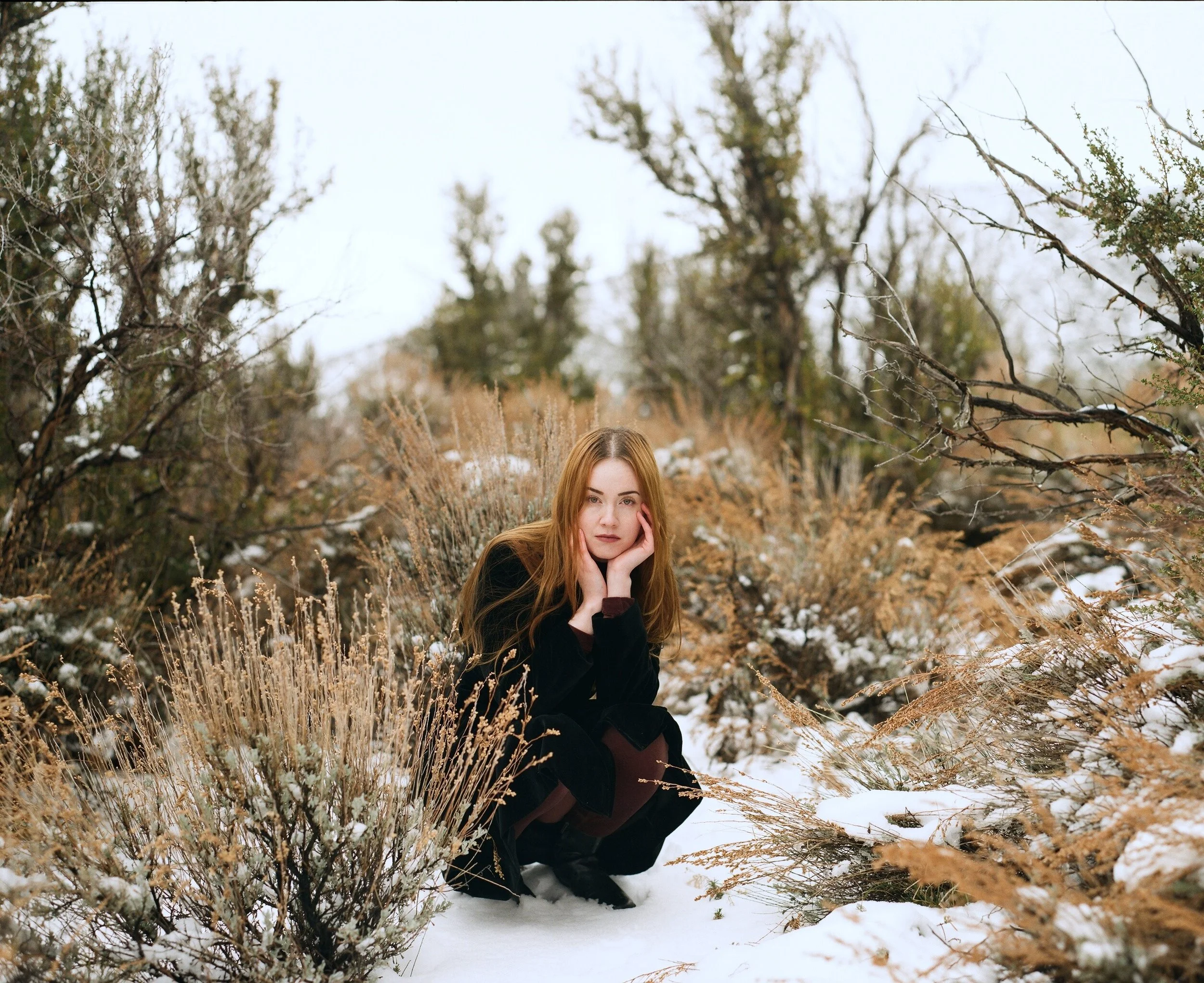 A young woman with long reddish hair, wearing a black jacket, sitting outdoors in a snowy natural setting with dry grass and leafless trees.