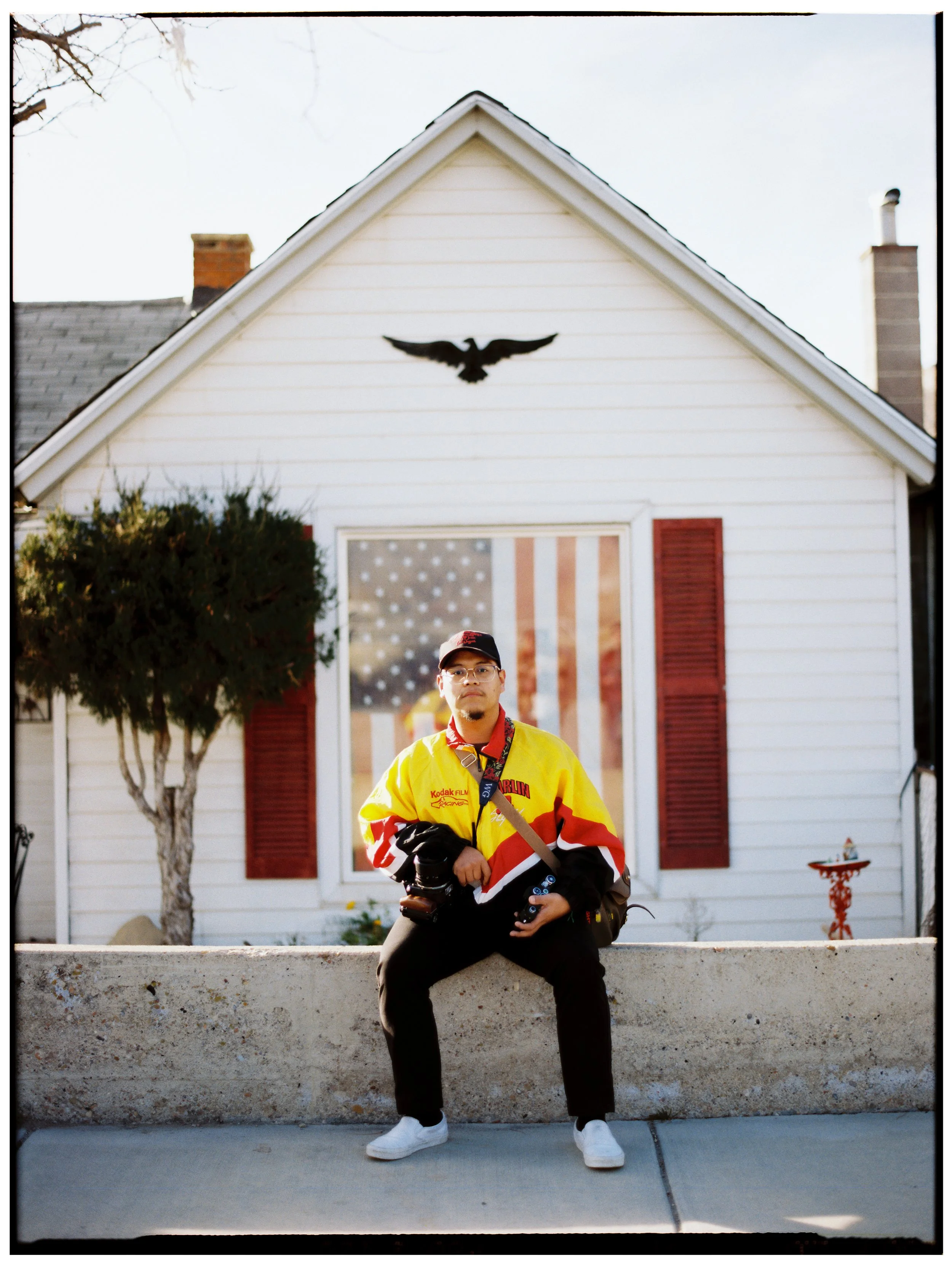 A young man wearing glasses, a yellow and red jacket, and black pants sitting on a concrete ledge in front of a white house with red shutters, an American flag window decoration, and an eagle decoration on the house's gable.