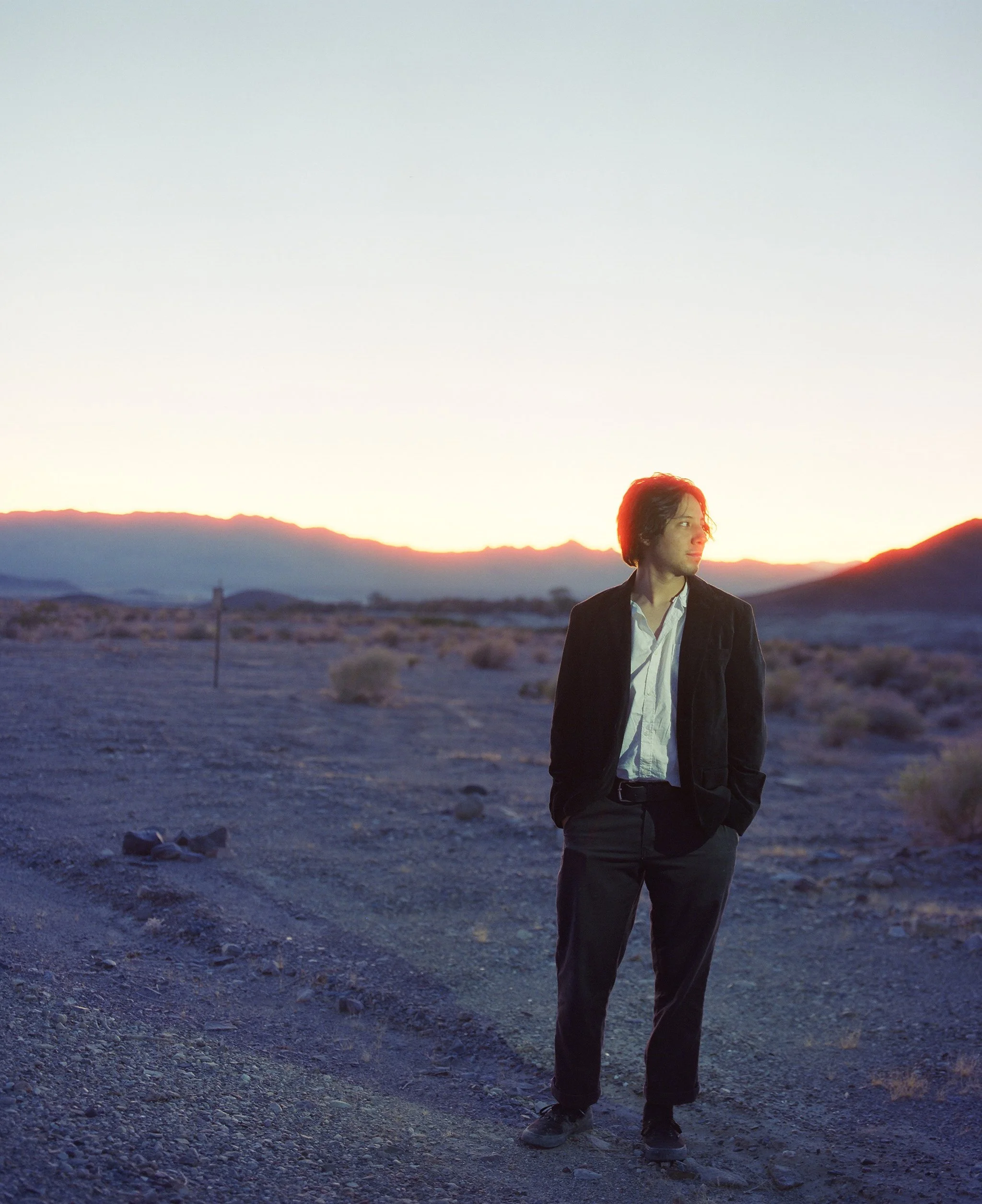 A person standing outdoors on a rocky landscape during sunset or sunrise, with mountains in the background and wearing a black jacket, white shirt, and black pants.