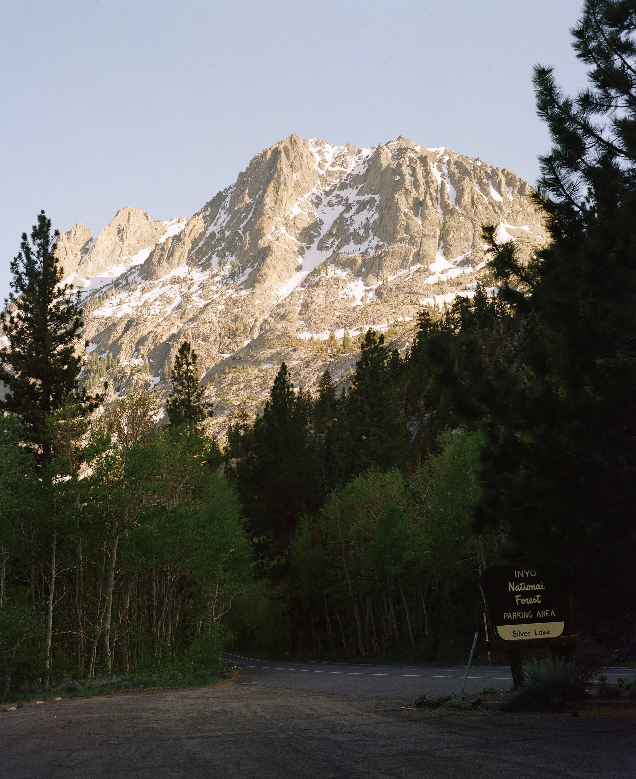 A mountainous landscape with snow-capped peaks and a forest of green trees in the foreground. A sign indicates the location as part of the Inyo National Forest, near Silver Lake, with a designated parking area.