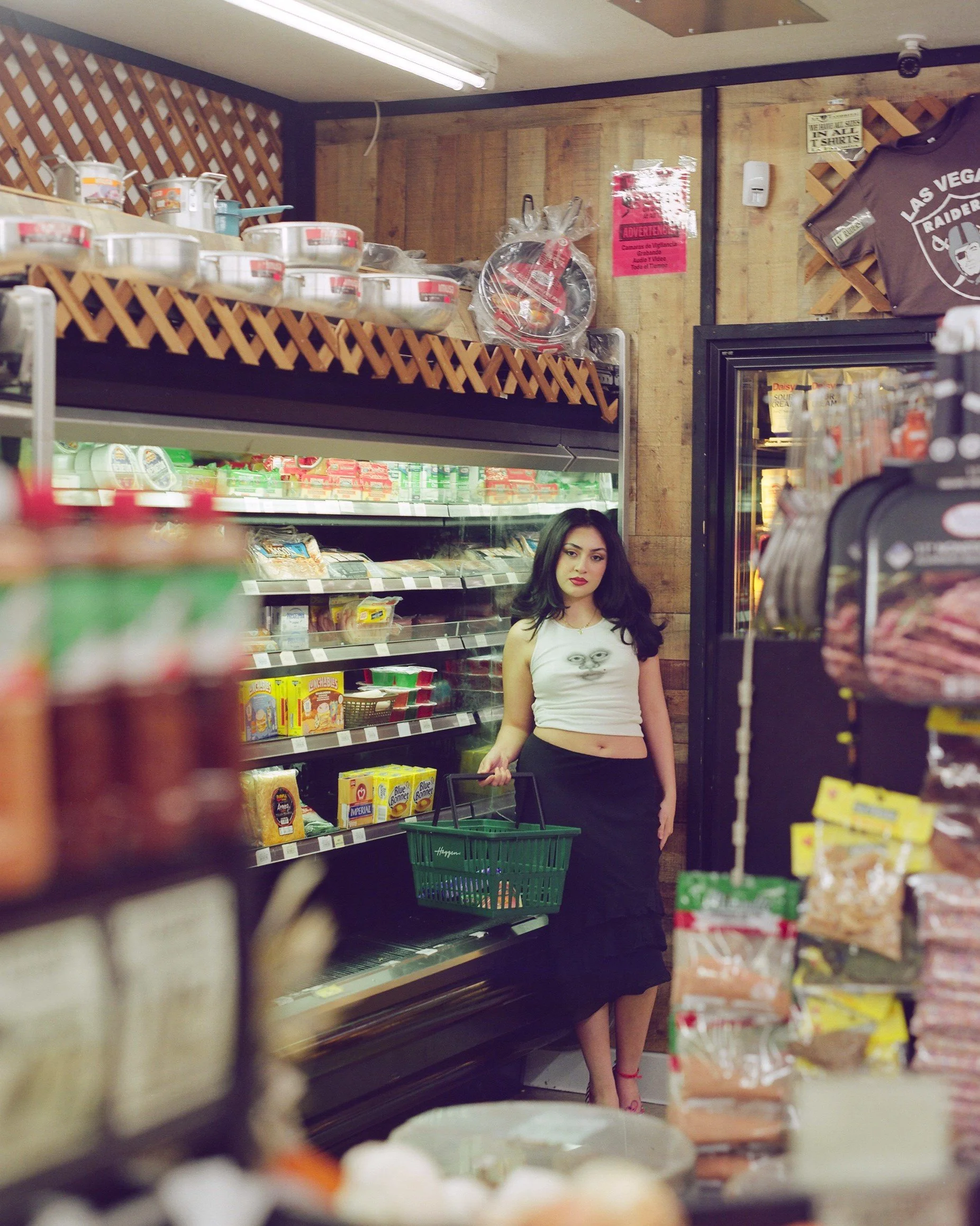 A woman with long dark hair, wearing a white crop top and a black skirt, is standing in a grocery store aisle holding a shopping basket. Shelves around her are stocked with various food products, and there is a display with snacks or candy in the for