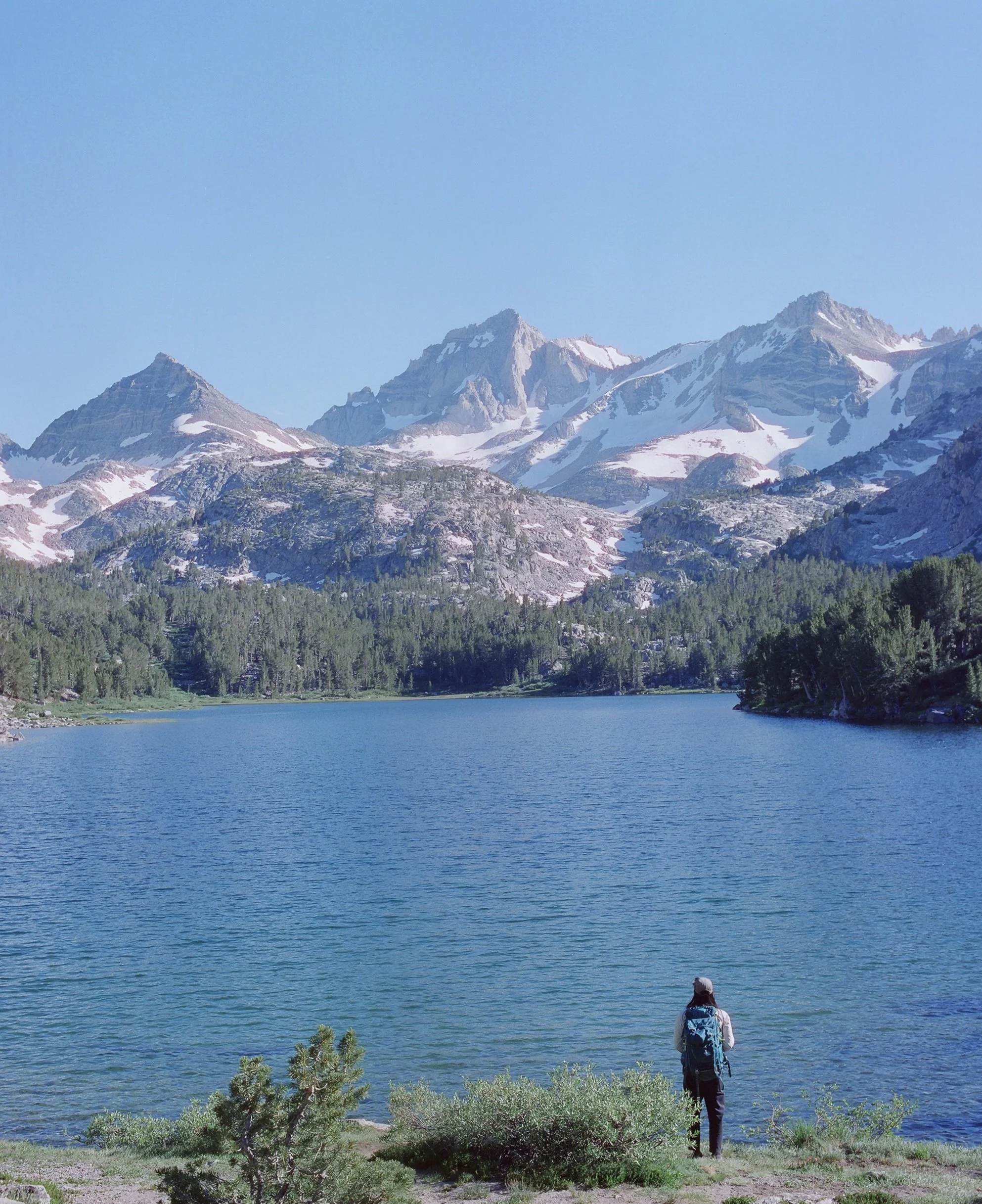 A person wearing outdoor gear with a backpack, standing near a lake surrounded by trees, with snow-capped mountains in the background under a clear blue sky.