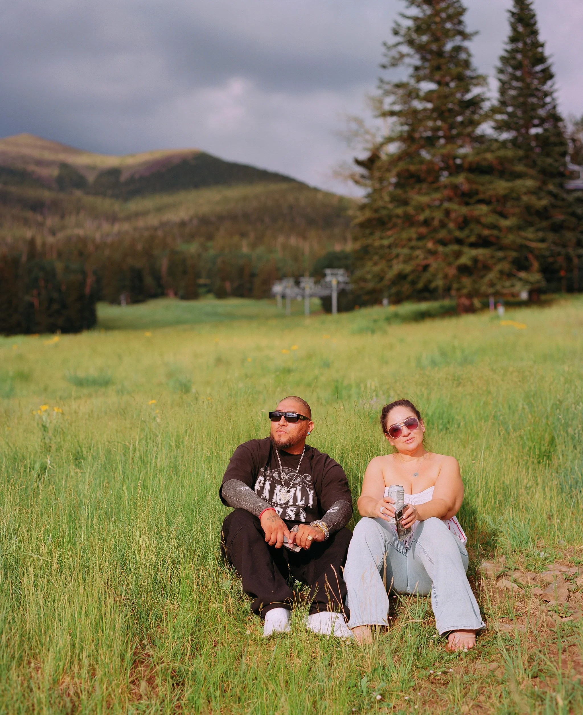 Two people sitting on grass in a field with trees and hills in the background, one man and one woman, both wearing sunglasses.