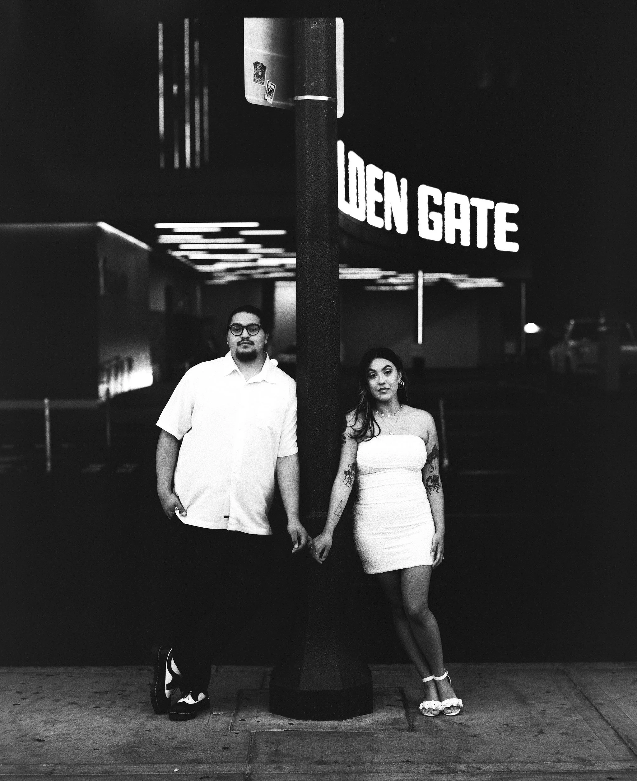 A black-and-white photo of a man and a woman standing in front of a sign that says "GEN GATE." The man has glasses, a goatee, and is wearing a white shirt. The woman has long dark hair, tattoos on her arms, and is wearing a strapless white dress and 