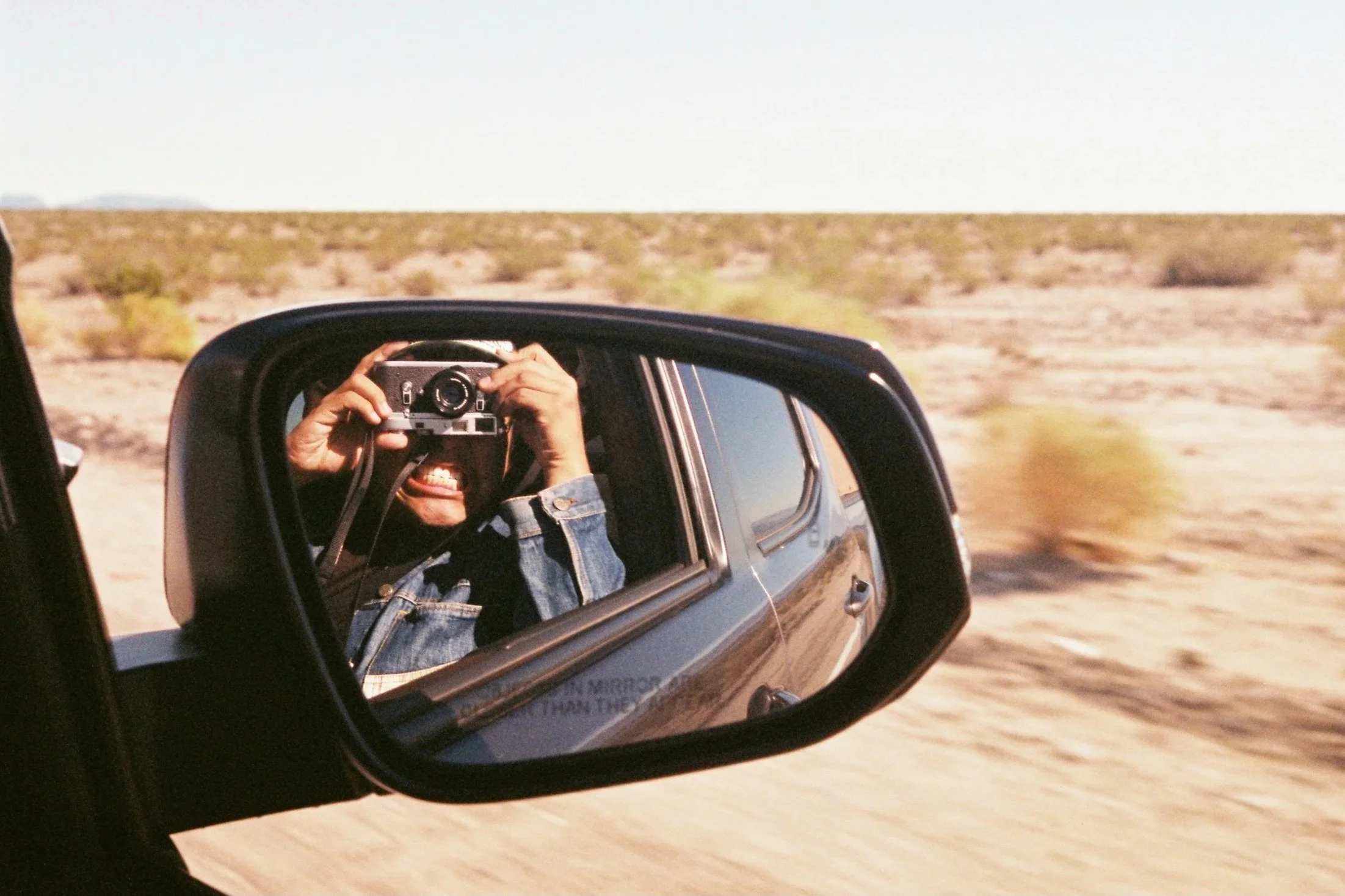 Person taking a photograph of themselves with a camera in the side mirror of a vehicle while driving through a desert landscape.