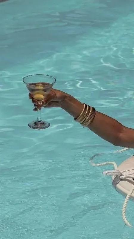 A person's arm extends out of a swimming pool, holding a cocktail glass with a lemon slice inside, while wearing multiple gold bangles.