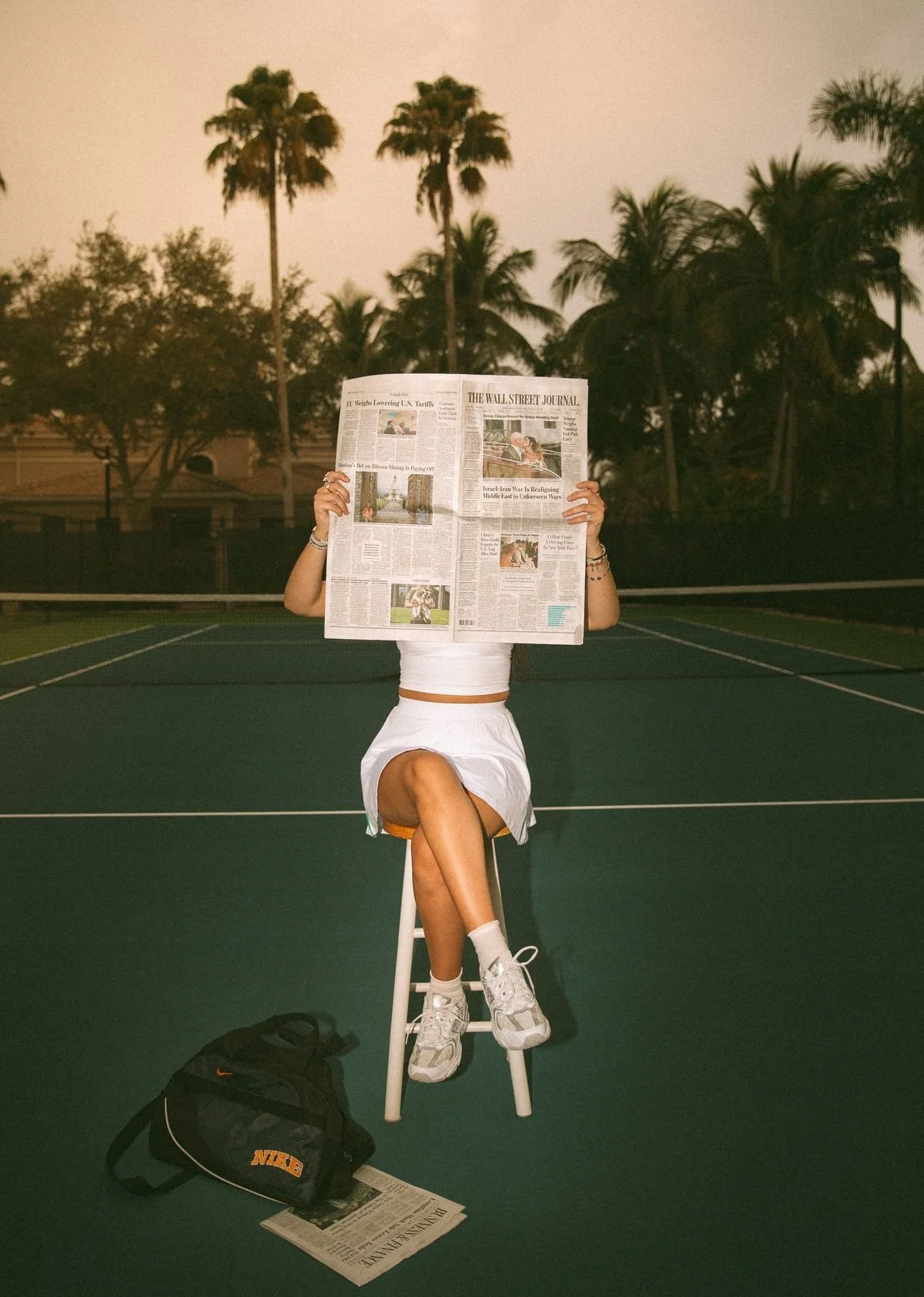 A person sitting on a stool on a tennis court reading a newspaper, with a backpack and a newspaper on the ground in front. The person is wearing a white outfit and sneakers. Trees and a building are in the background during dusk.