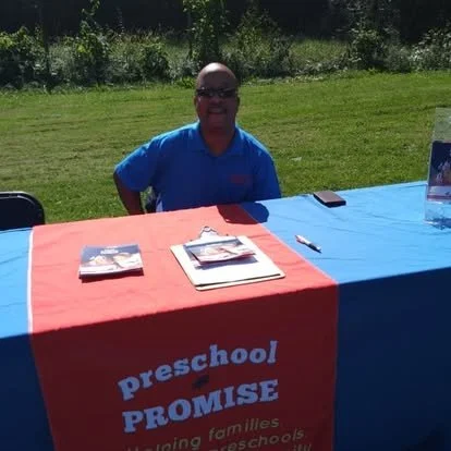 A woman sitting at a table outdoors with a red and blue tablecloth that reads 'Preschool Promise' on a sunny day.