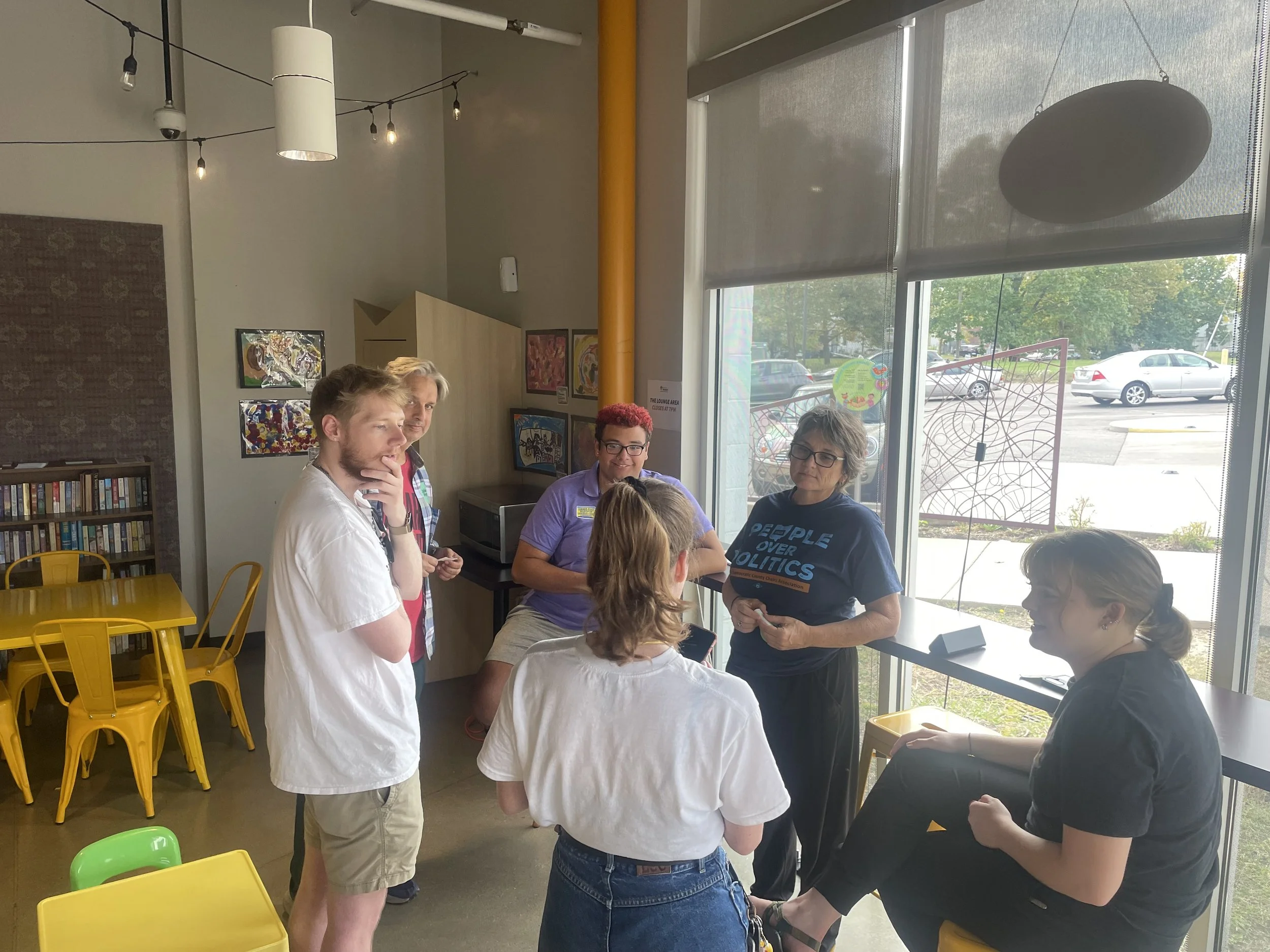 A group of six people having a discussion in a brightly lit room with large windows. Four women and two men are engaging in conversation, with some sitting and some standing. The room has yellow chairs, artwork on the walls, and a bookshelf. Outside the window, parked cars and trees are visible.