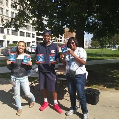 Three people standing outdoors near a park, holding informational pamphlets or flyers, with trees and a building in the background.