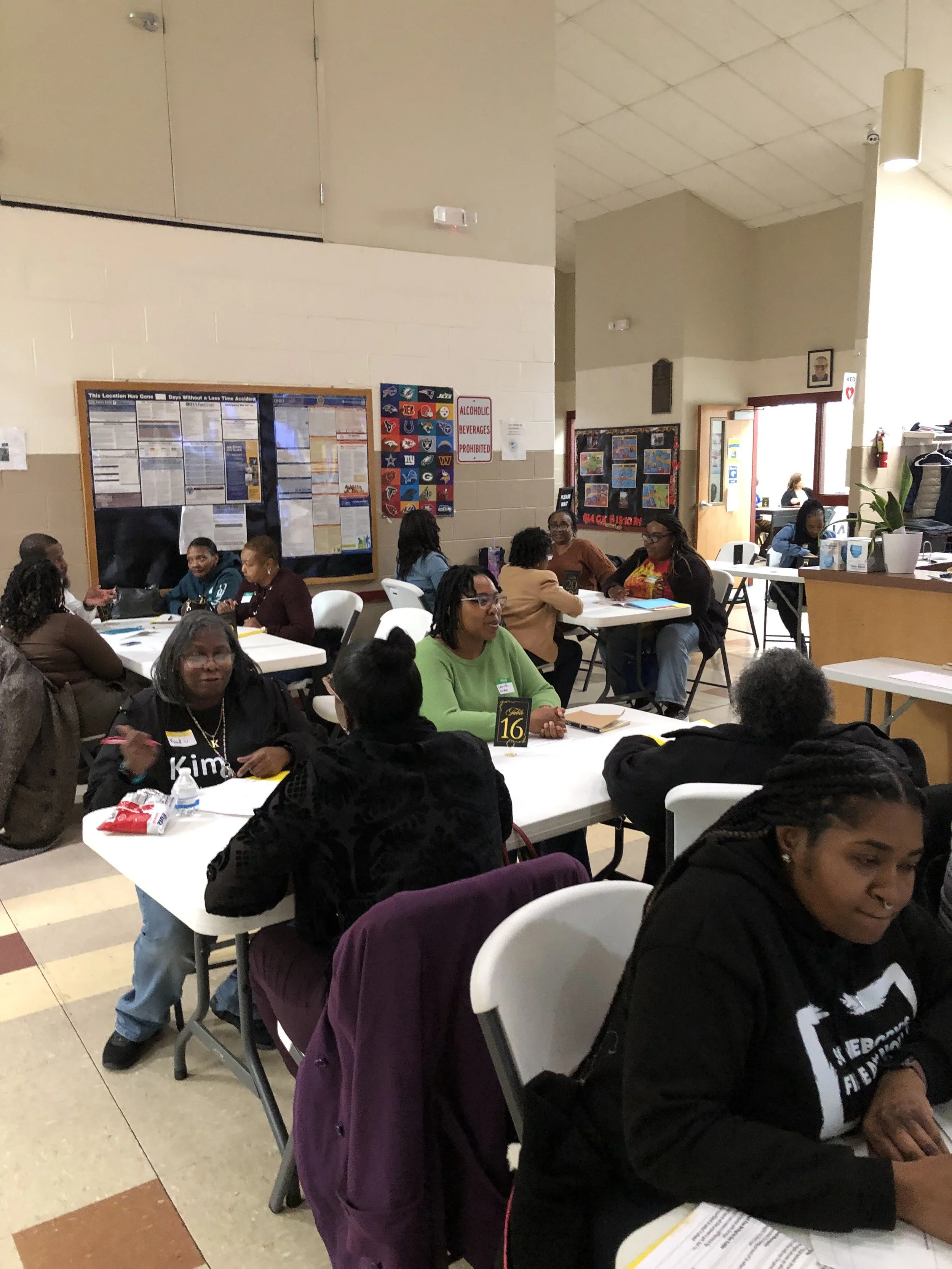 A group of diverse women sitting at tables in a room, engaged in conversation. The room has bulletin boards on the walls, some with colorful posters and notices. There are tables numbered with small signs, and the setting appears to be a community or meeting space.