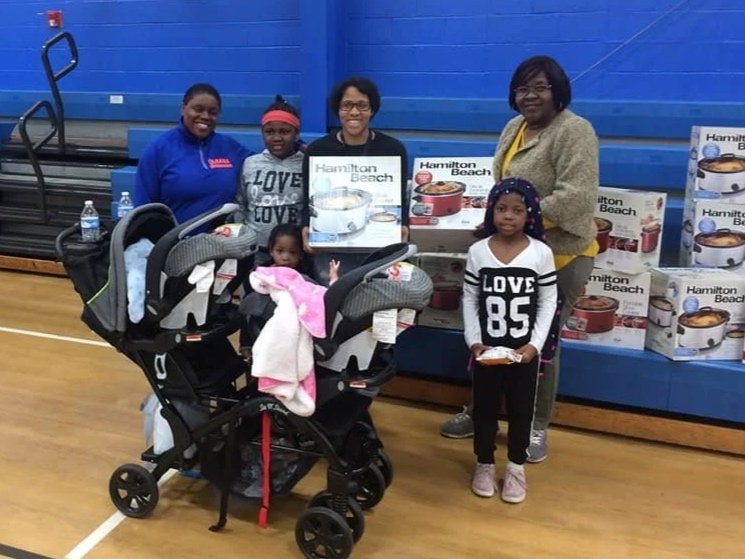 A group of children and adults standing behind strollers and boxes of Hamilton Beach kitchen appliances in a gymnasium.
