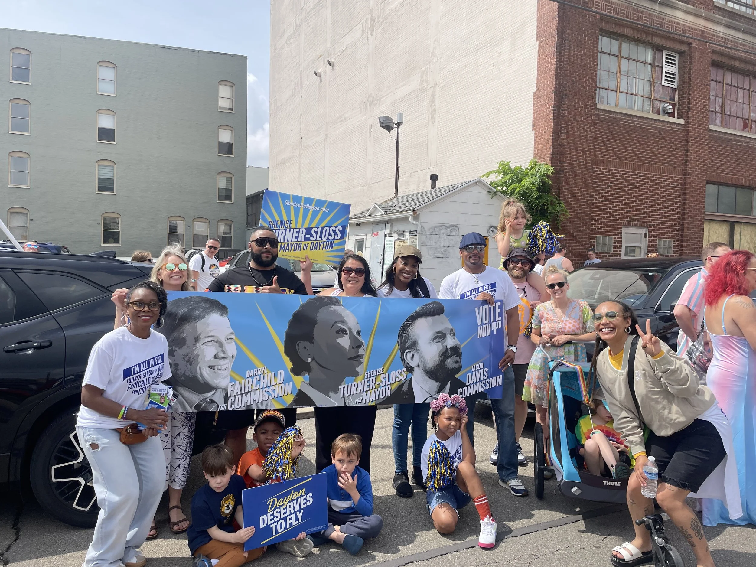 Group of people participating in a parade or rally holding campaign signs and banners for local elections in Dayton, Ohio, with children and adults smiling and making peace signs, some dressed casually, some with colorful outfits, in an urban setting with cars and buildings in the background.