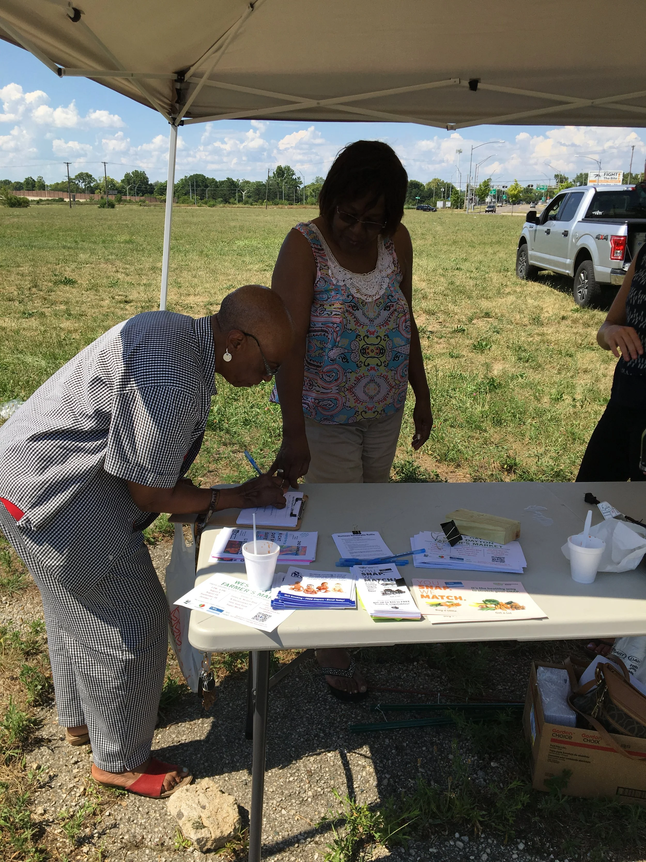 Two women at an outdoor table under a canopy, filling out paperwork and talking. The table has flyers, forms, and cups on it.