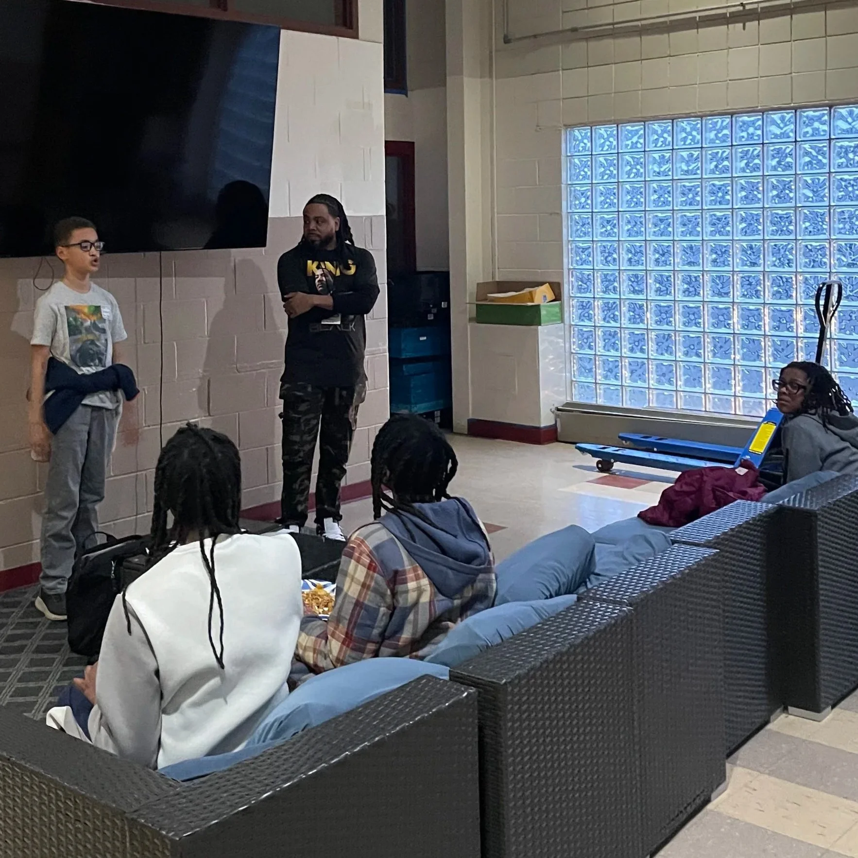 A group of five people in a room with a large TV, glass block window, and a gray couch. Two individuals are standing near the TV, one young boy with glasses and another adult with long dreadlocks. Three others are seated on the couch, watching the standing pair. One person on the right side is looking in their direction, with a skateboard on the floor nearby.