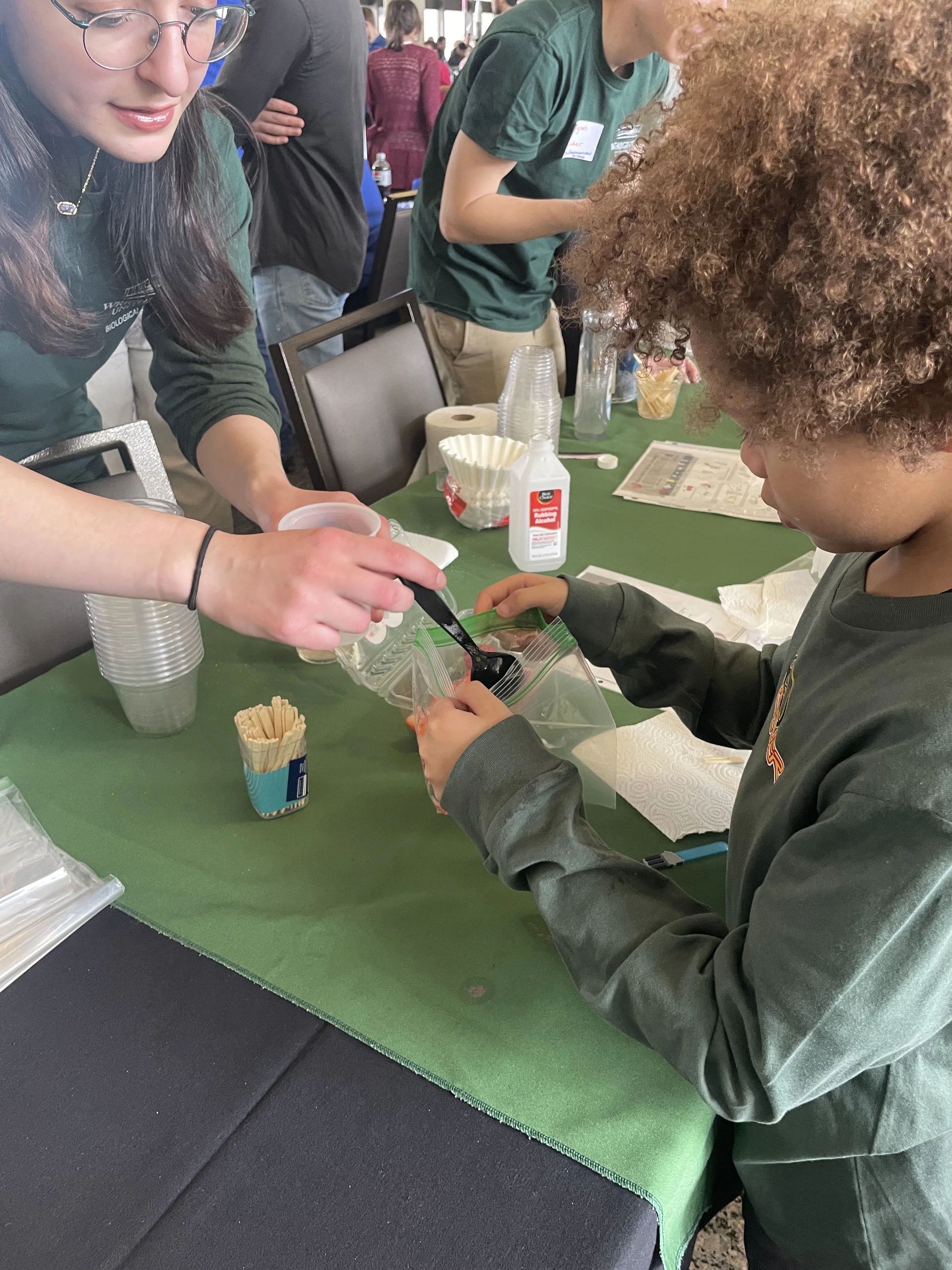 A young boy with curly hair getting a scoop of food from a woman at an event with a green tablecloth, surrounded by cups, napkins, and condiments.