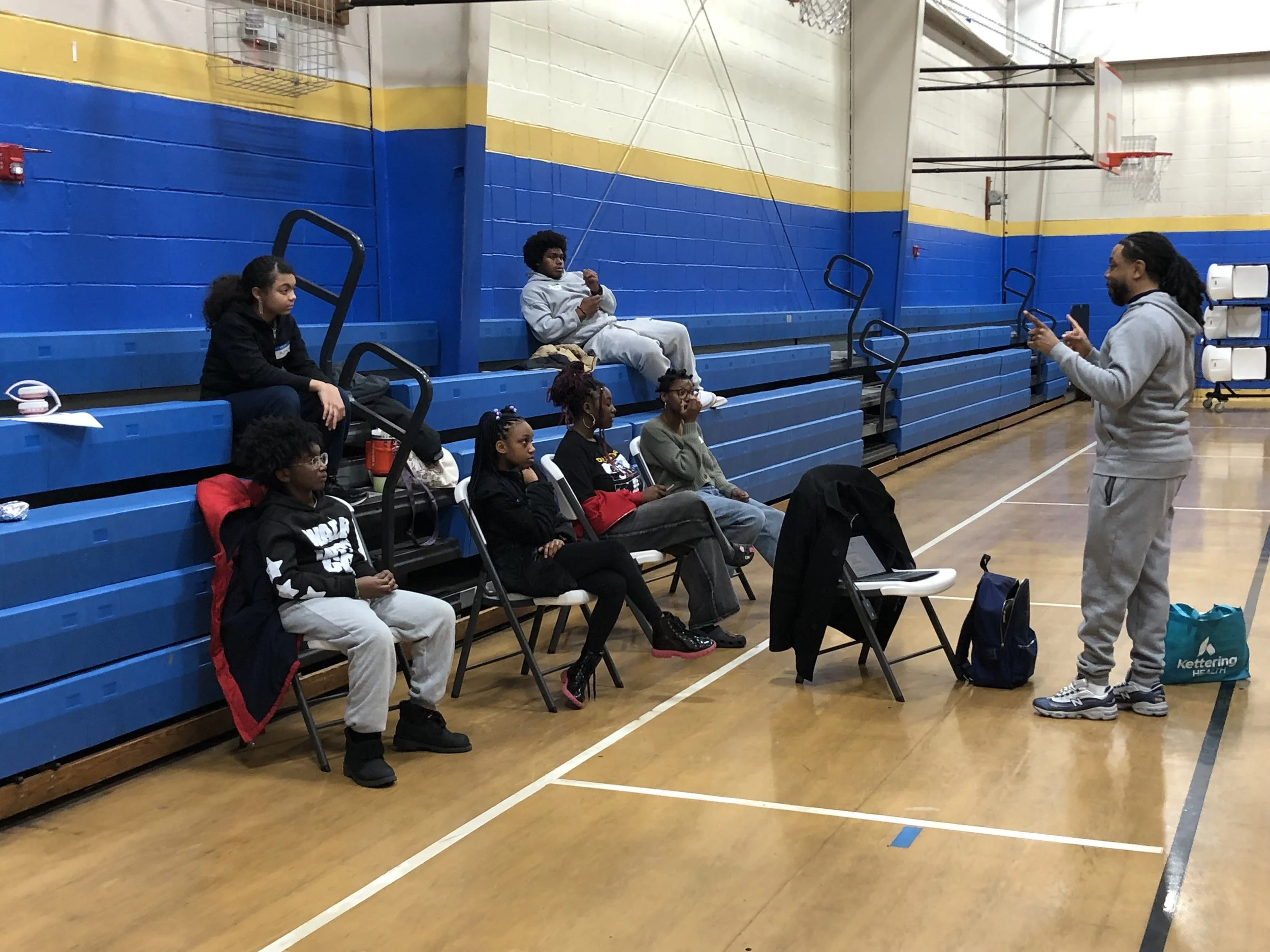A group of children and a man sitting on chairs and bleachers in a gymnasium. The man is standing and appears to be speaking or giving a presentation to the children.