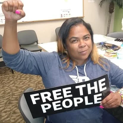 A woman sitting at a table holding a sign that says 'Free the People' and raising her fist.