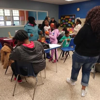 Children and adults gathered in a classroom with tables, chairs, and a bulletin board decorated with colorful letters.