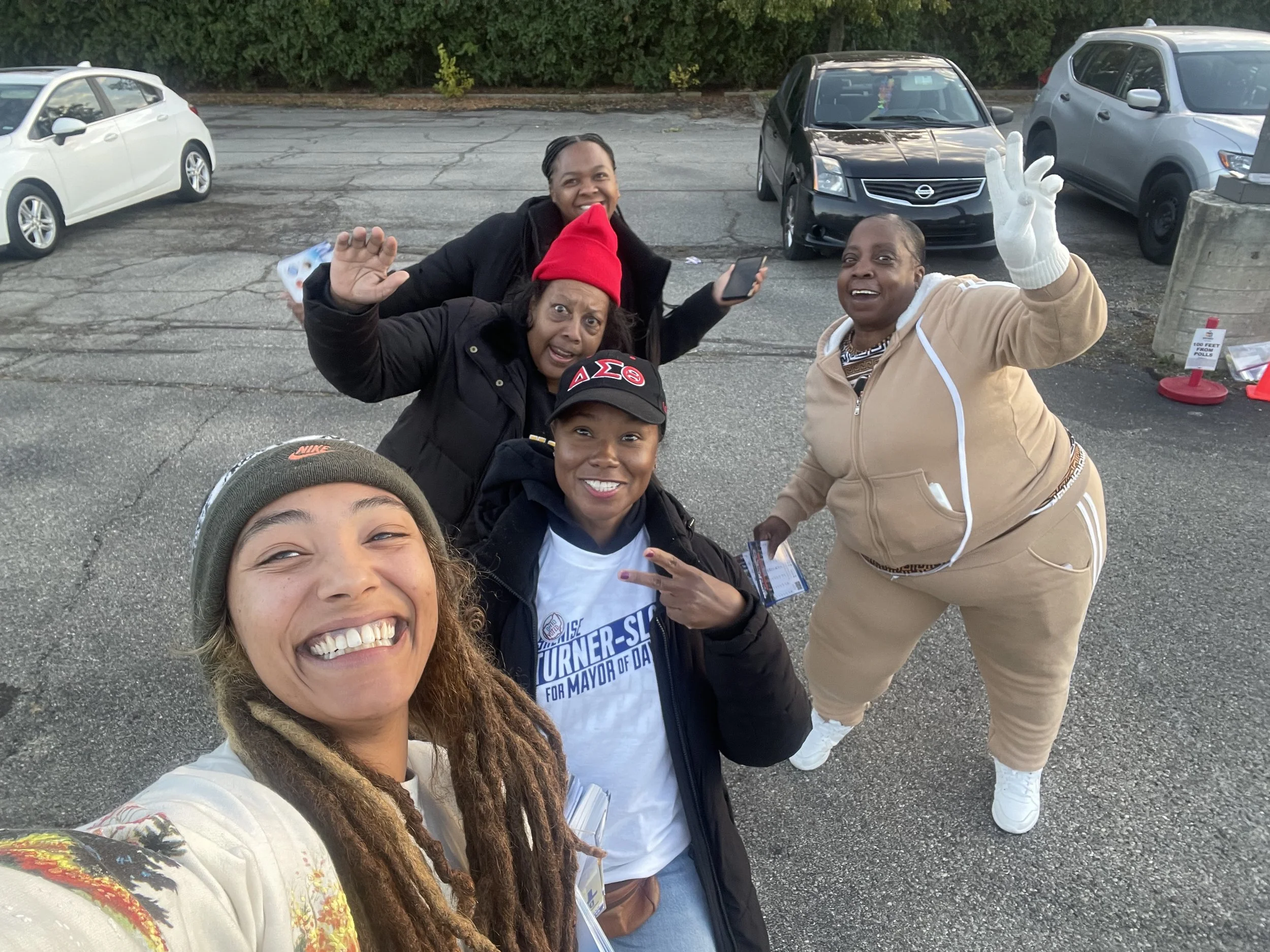 Group of five smiling women taking a selfie in a parking lot, some waving and making peace signs, with cars in the background.