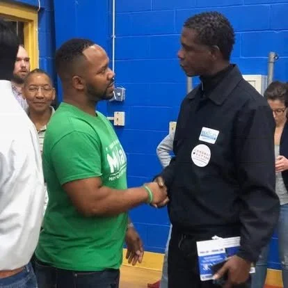 Two men shaking hands, one wearing a green shirt and the other in a black shirt, in a room with blue walls and people observing.