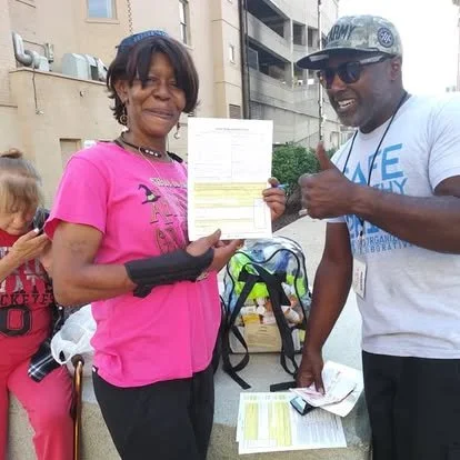 A woman holding a document and smiling, standing next to a man giving a thumbs-up, outdoors in an urban setting.