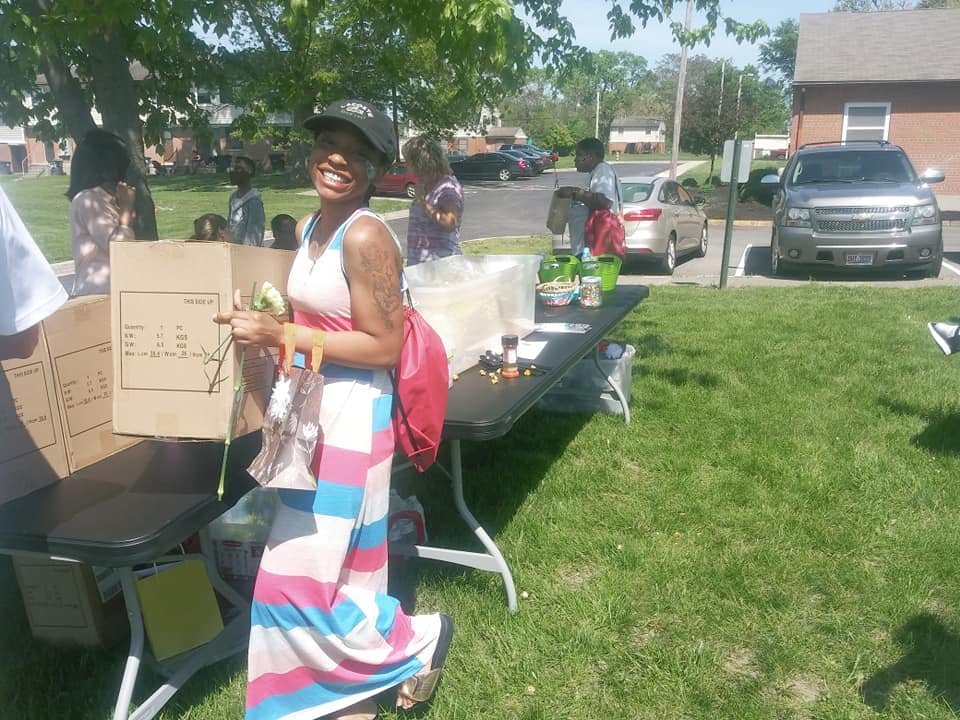 Smiling woman with tattoos and a black cap at outdoor community event, standing at a table with food and drink supplies, surrounded by people and parked cars on a sunny day.