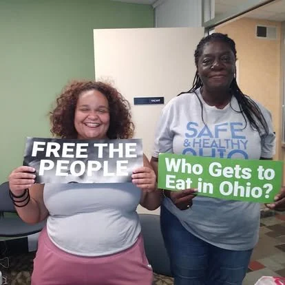 Two women smiling, holding signs that say 'Free the People' and 'Who Gets to Eat in Ohio?' standing indoors against a green wall.